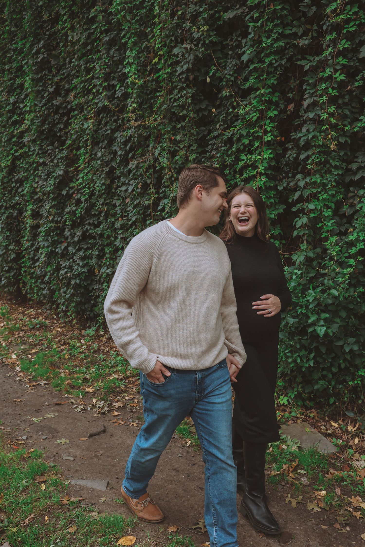 A smiling couple walks along a leaf-strewn path beside a wall of green ivy, the woman in a black dress laughing as the man looks on.