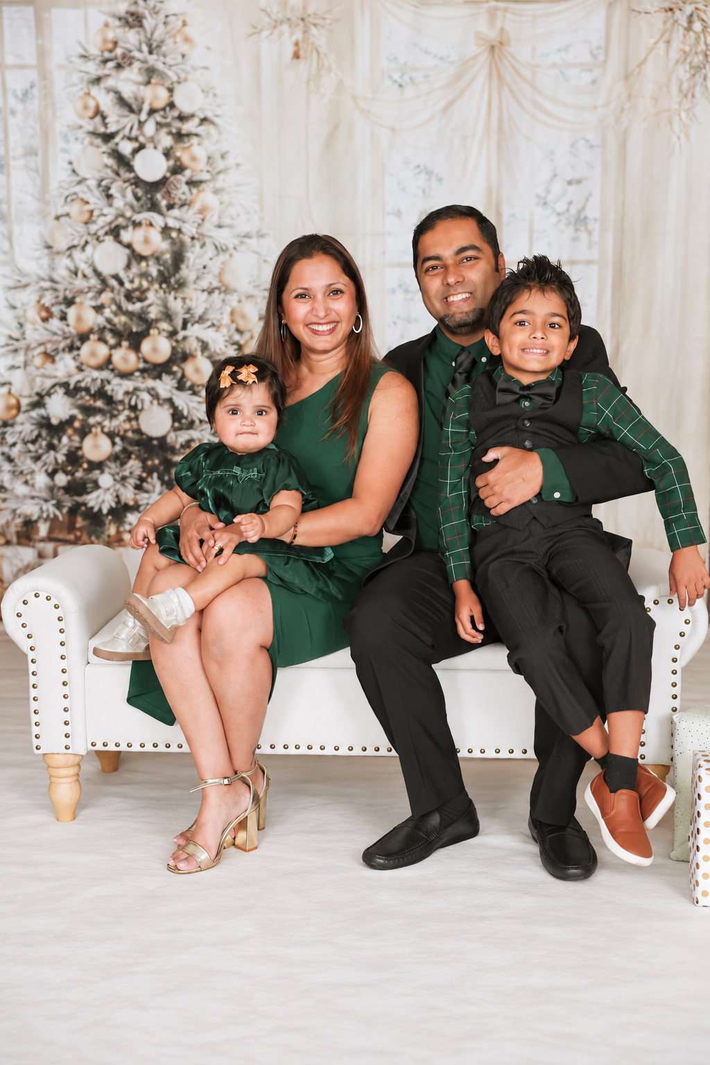 Family of four in matching green outfits posing on white sofa before decorated Christmas tree.