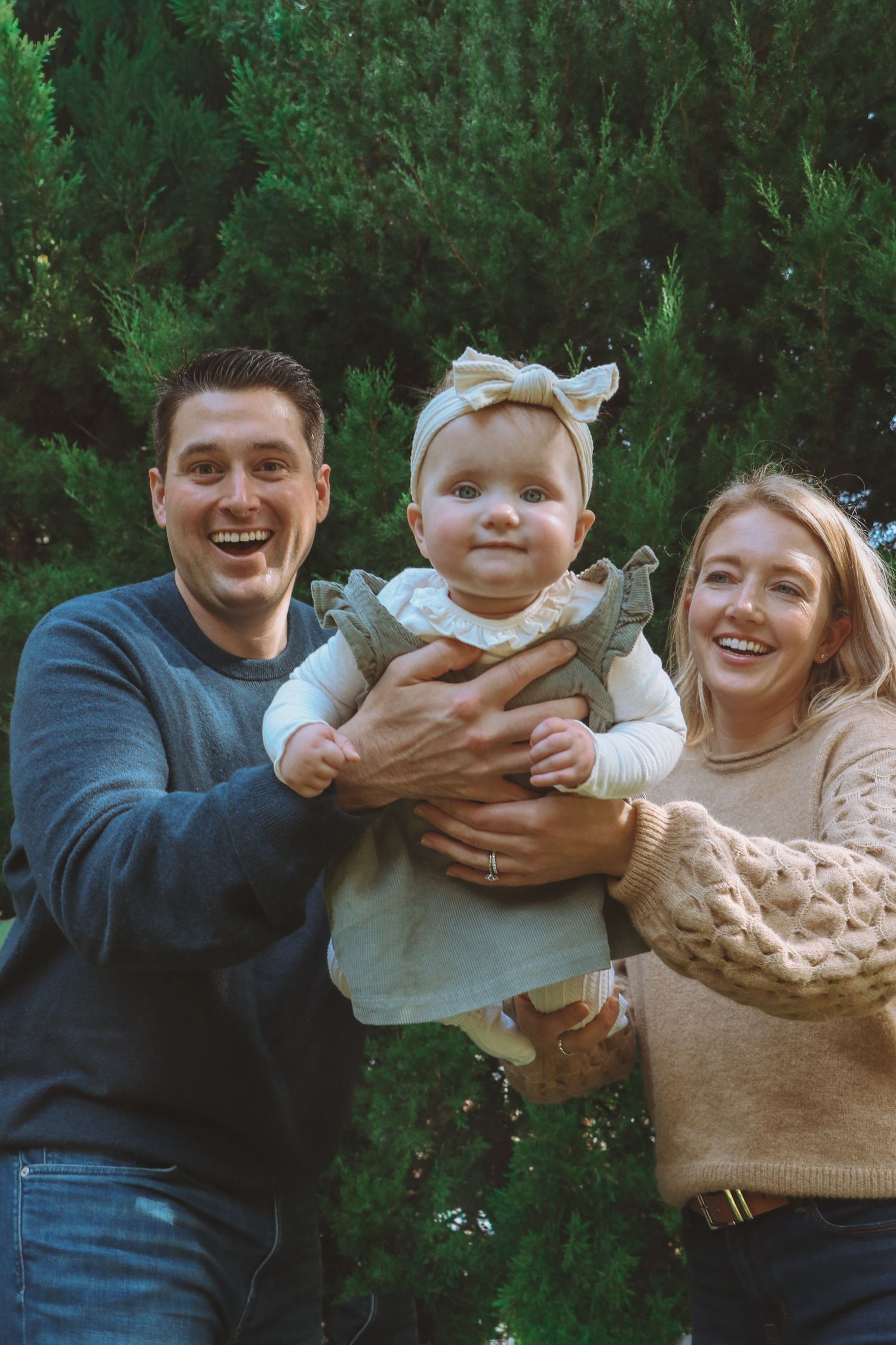 Smiling parents hold their wide-eyed baby girl in a green dress in front of tall evergreen trees.
