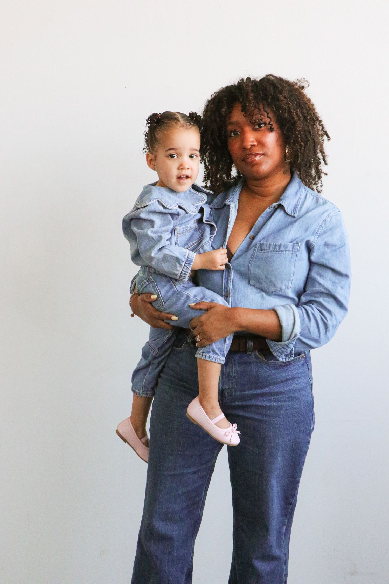 Black mother in denim outfit holding toddler daughter in matching denim against white wall.