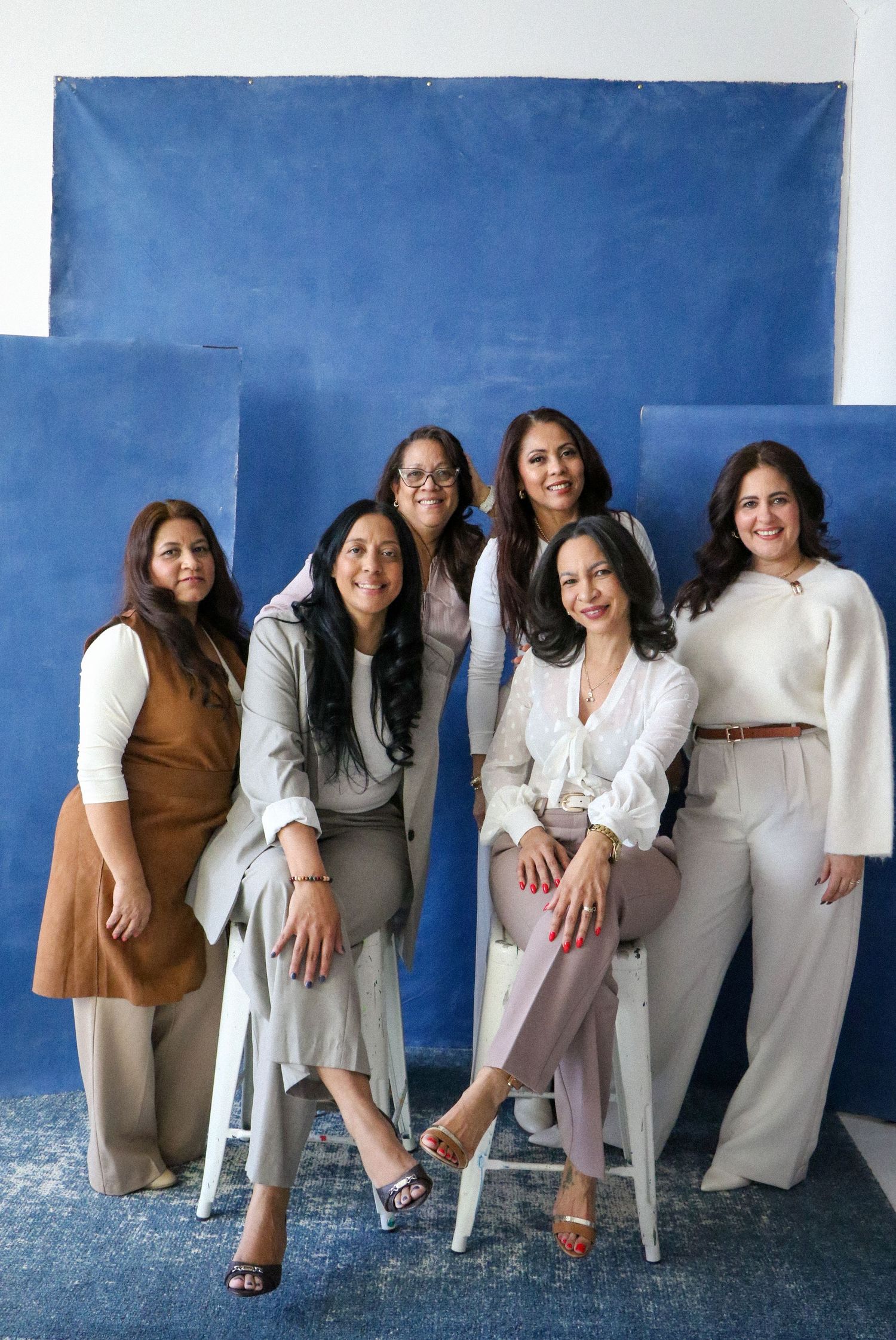 Six diverse women posing together in neutral tones against a blue painted backdrop in a studio.