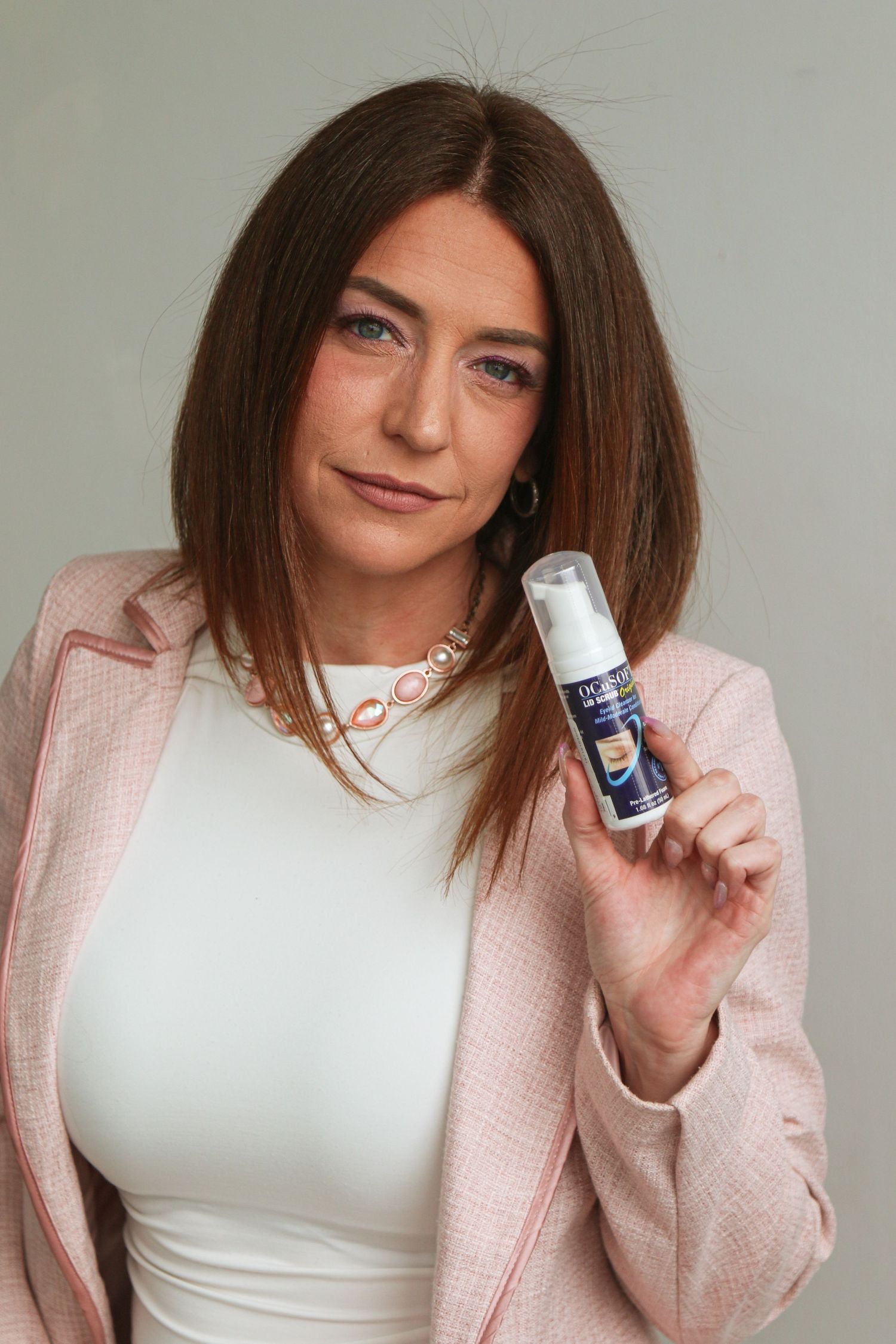 Woman in pink blazer holding a skincare product bottle against a neutral gray background.