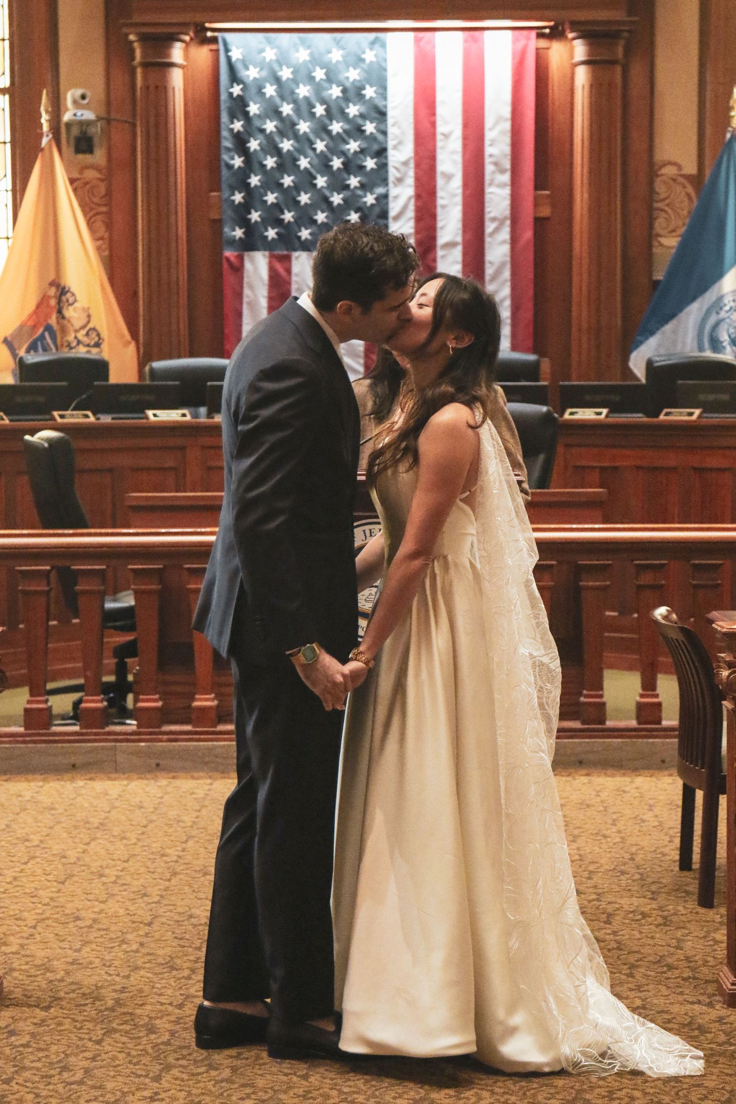 Couple sharing first kiss as newlyweds in a courthouse ceremony with American flag backdrop.