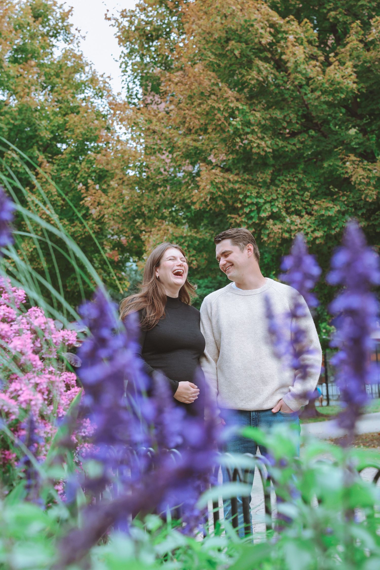 Laughing couple in casual sweaters stand together in a garden surrounded by purple and pink flowers with colorful autumn trees behind them.