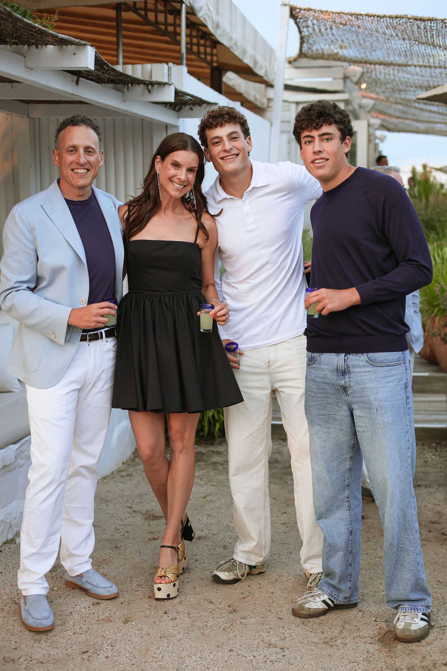 Four people in casual summer attire pose together outdoors on a sandy path near a beachside building at dusk, holding drinks.