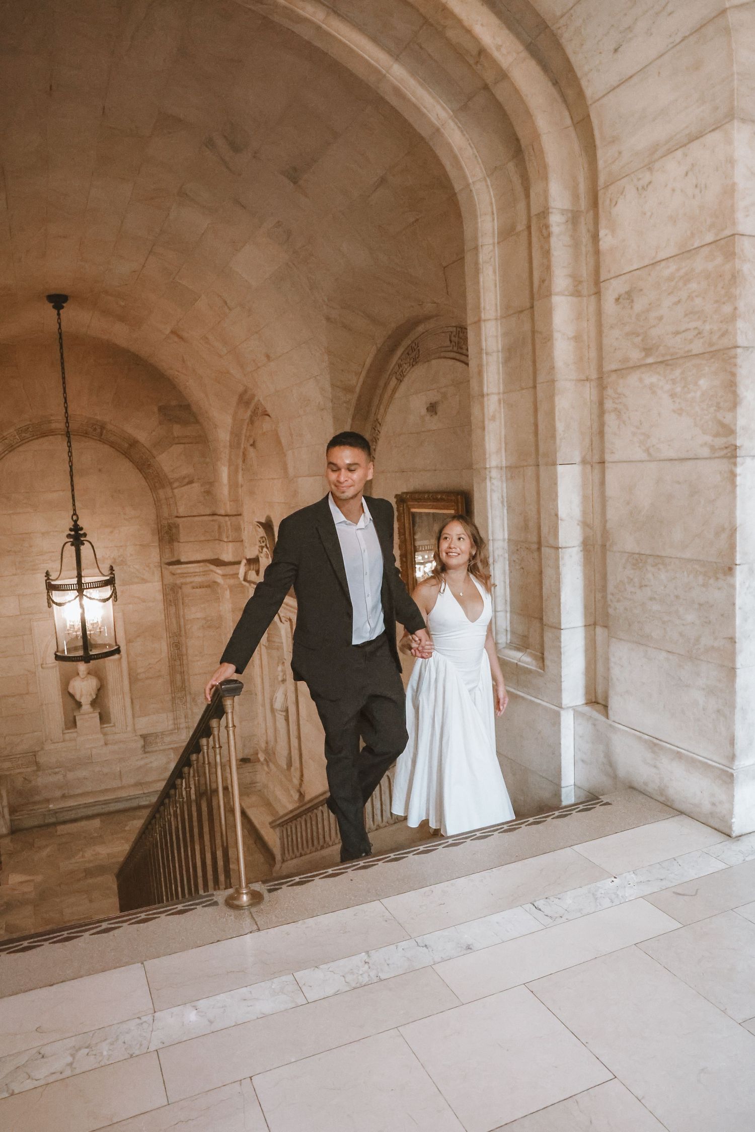 Couple in a black suit and white dress hold hands while climbing a marble staircase in a grand stone building with arched ceilings.