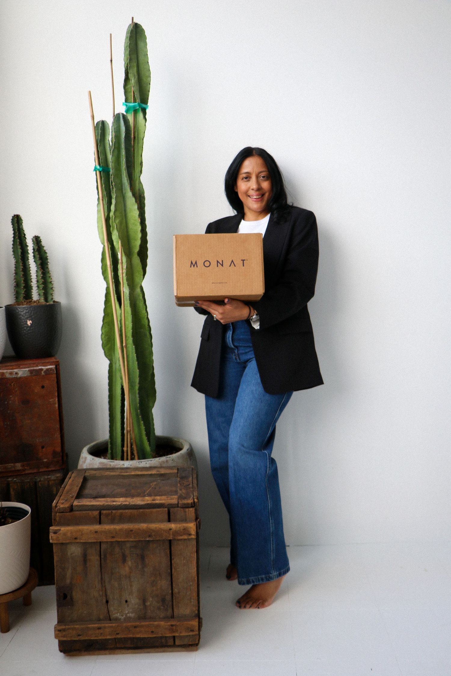 Smiling woman holding MONAT box standing beside tall cactus in bright studio setting.