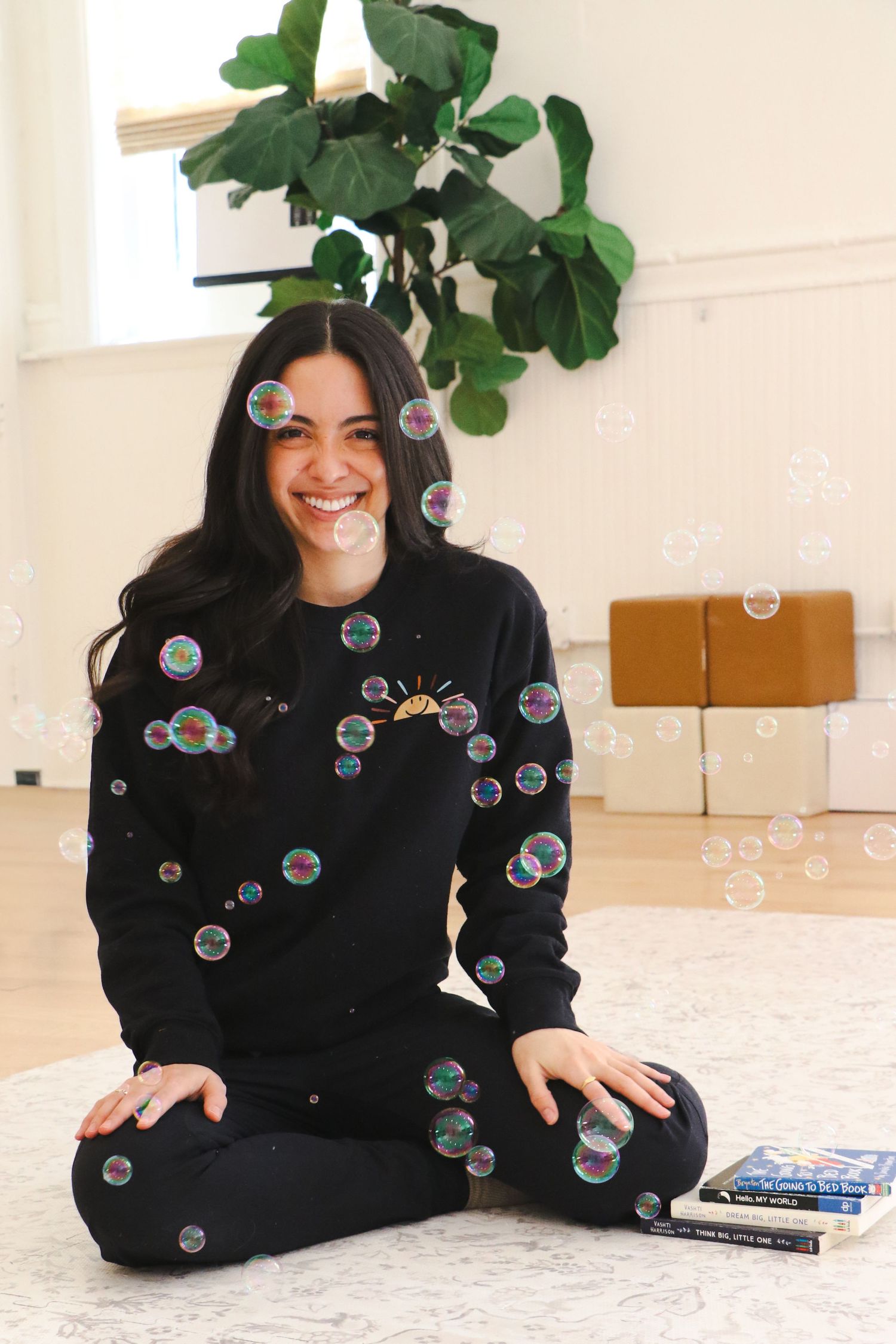 Happy young woman in black sweatshirt sitting cross-legged on floor surrounded by floating soap bubbles.