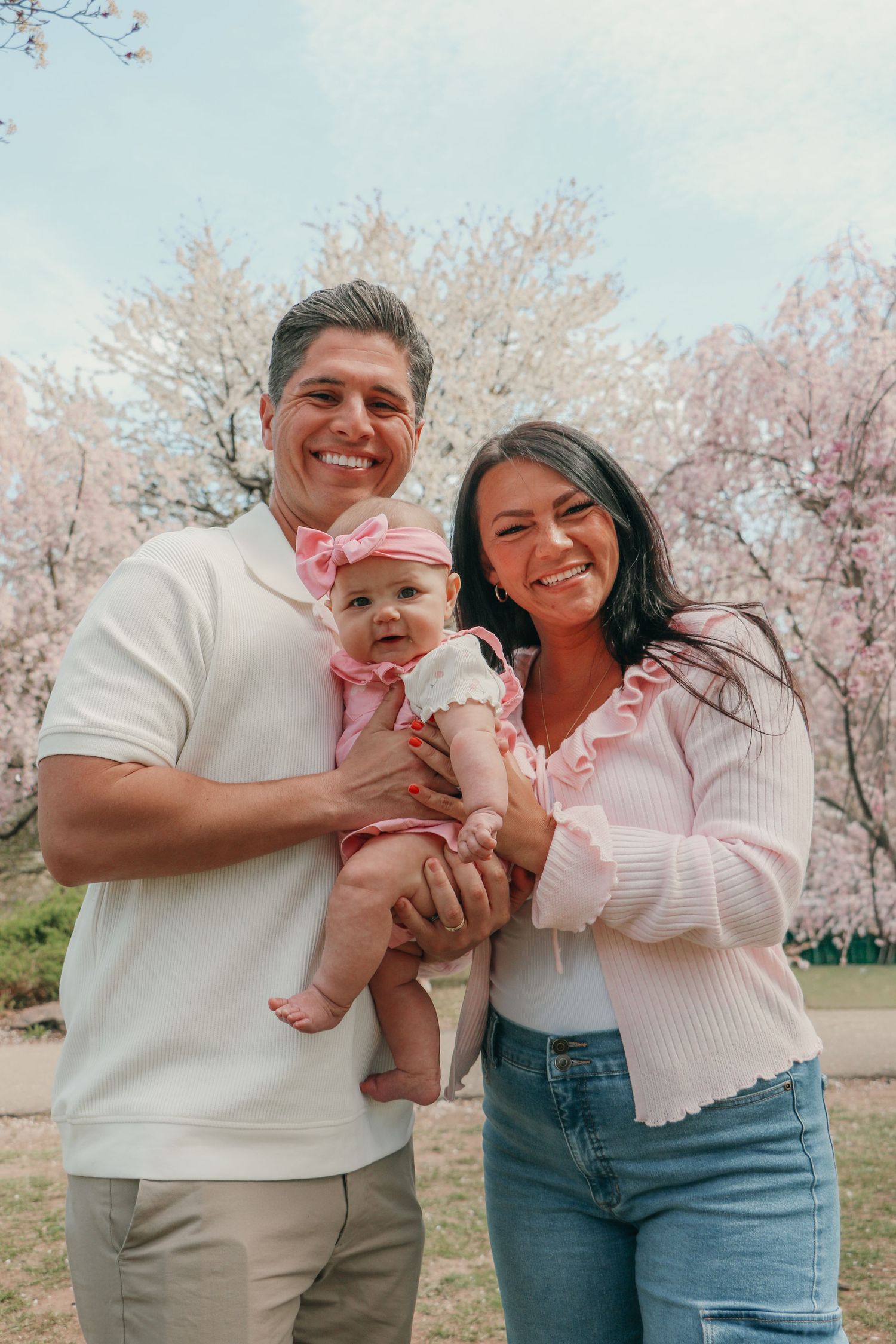 Young family with baby girl in pink bow poses among cherry blossoms in spring.