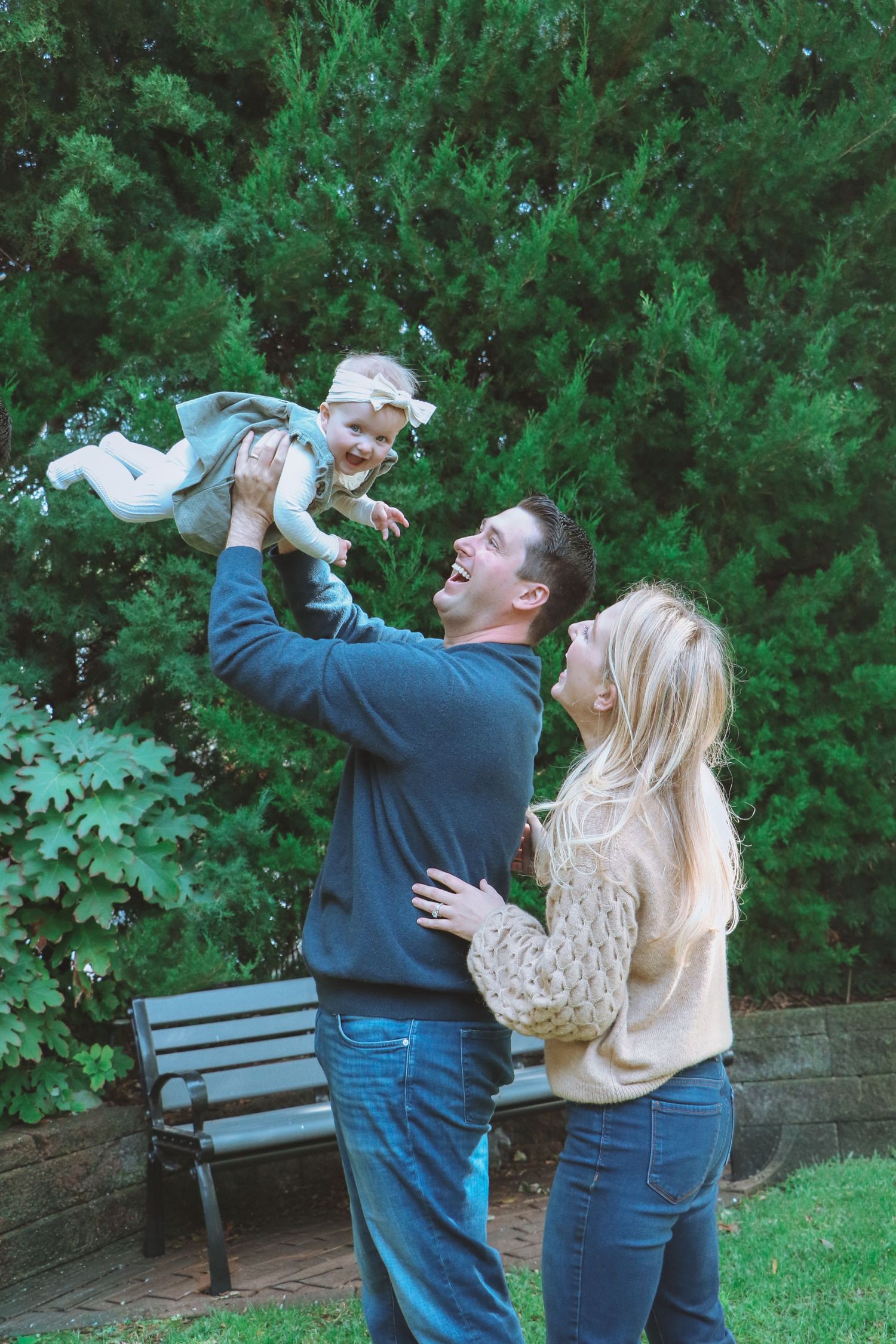 Joyful parents lift their giggling baby girl in the air in front of lush green evergreen trees.