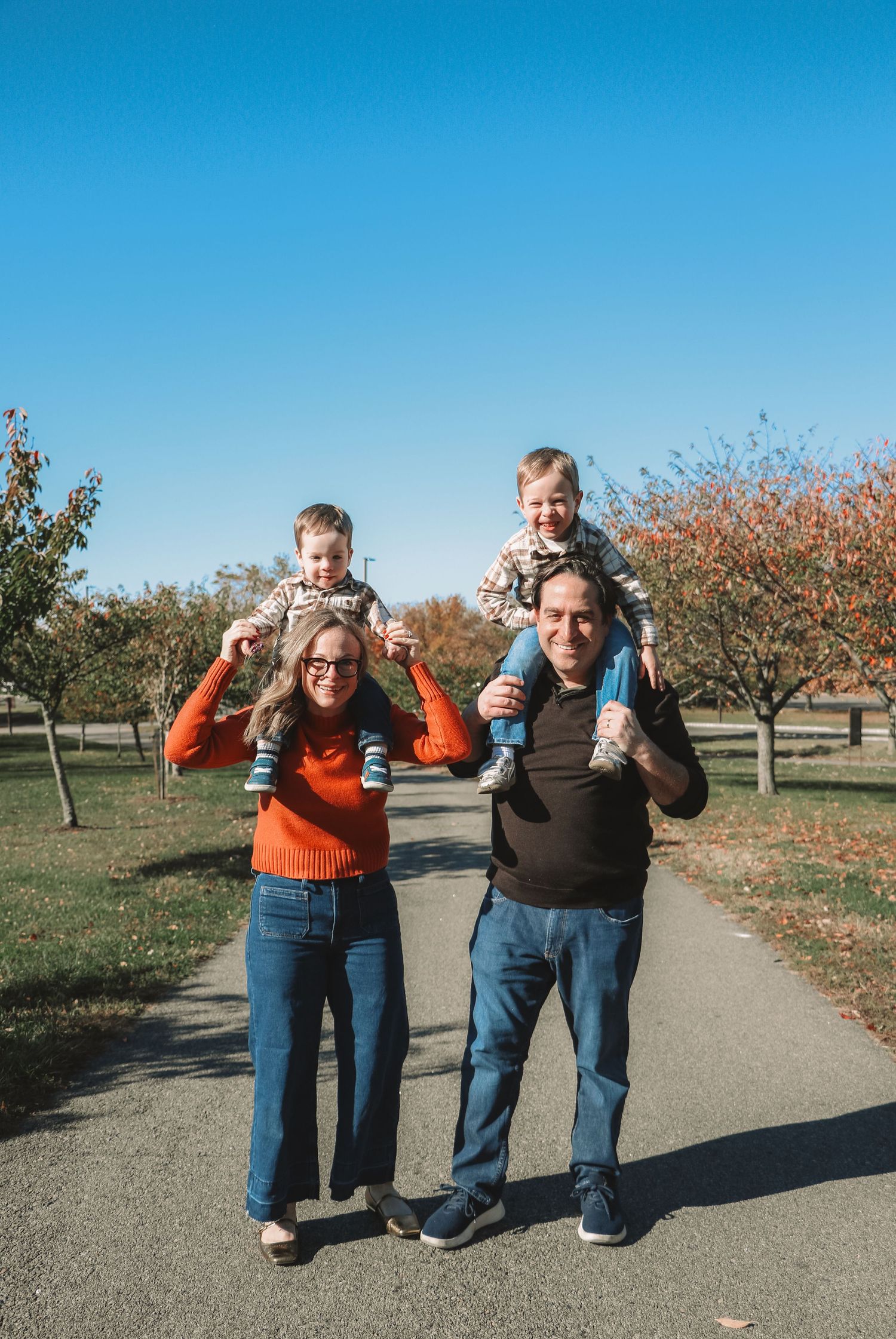 Smiling parents carrying twin toddlers on their shoulders on a sunny tree-lined park path in fall.