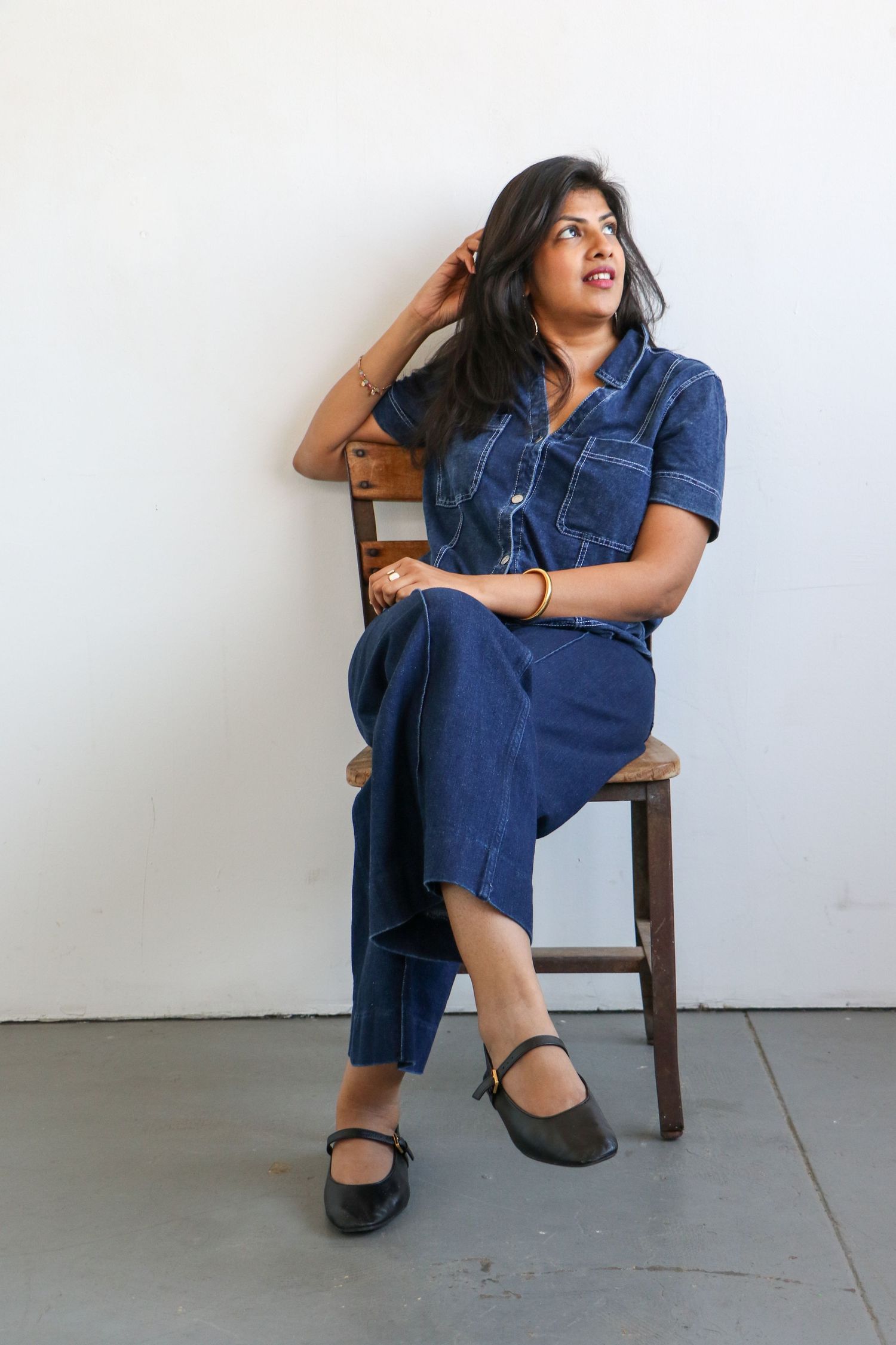 Woman in blue denim jumpsuit sitting on wooden chair, looking up thoughtfully against white wall.