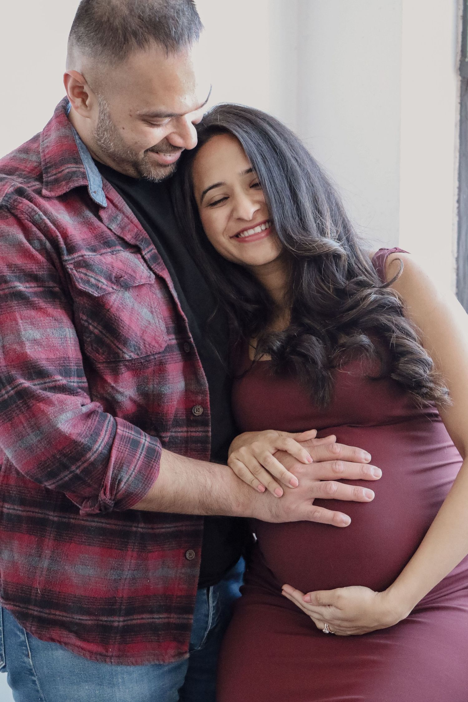 Expecting couple embracing by window, hands on pregnant belly, sharing tender moment together.
