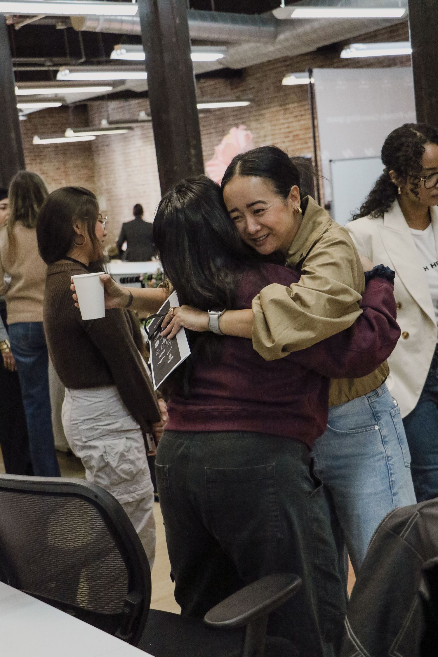 Two women hugging warmly at a casual indoor gathering, one holding an ultrasound photo.