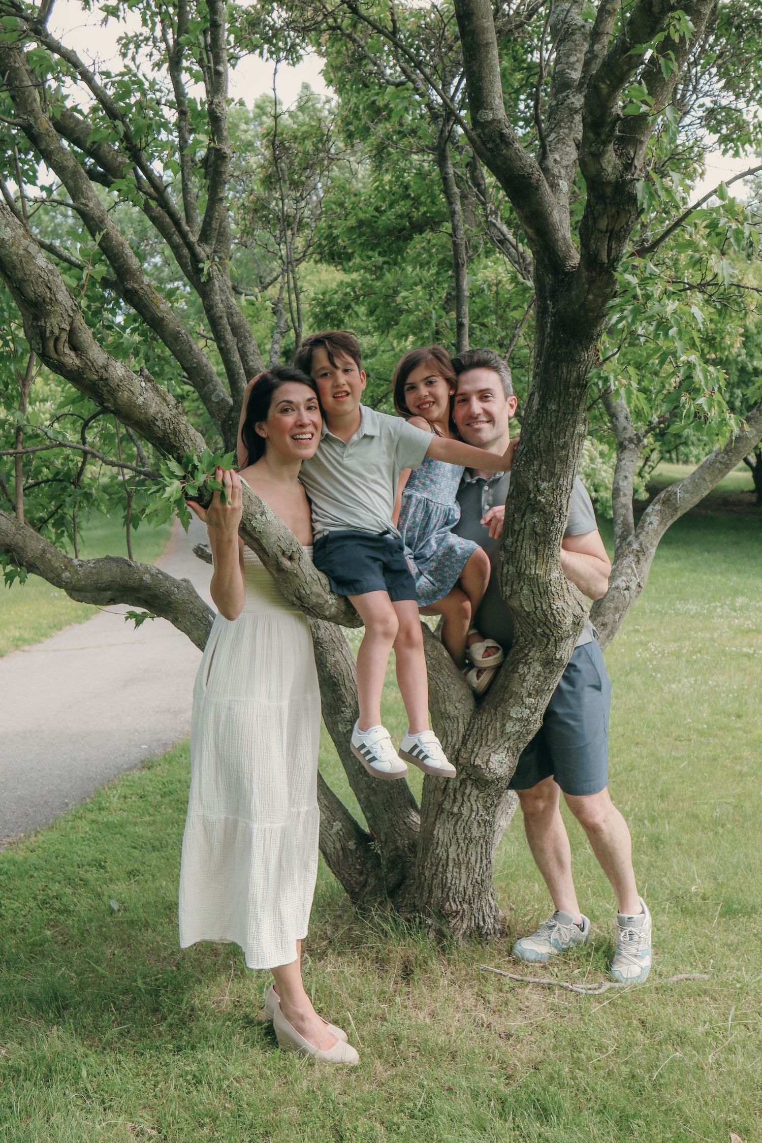 Happy family of four posing by a tree in a lush green park, parents smiling with two children sitting on branches.