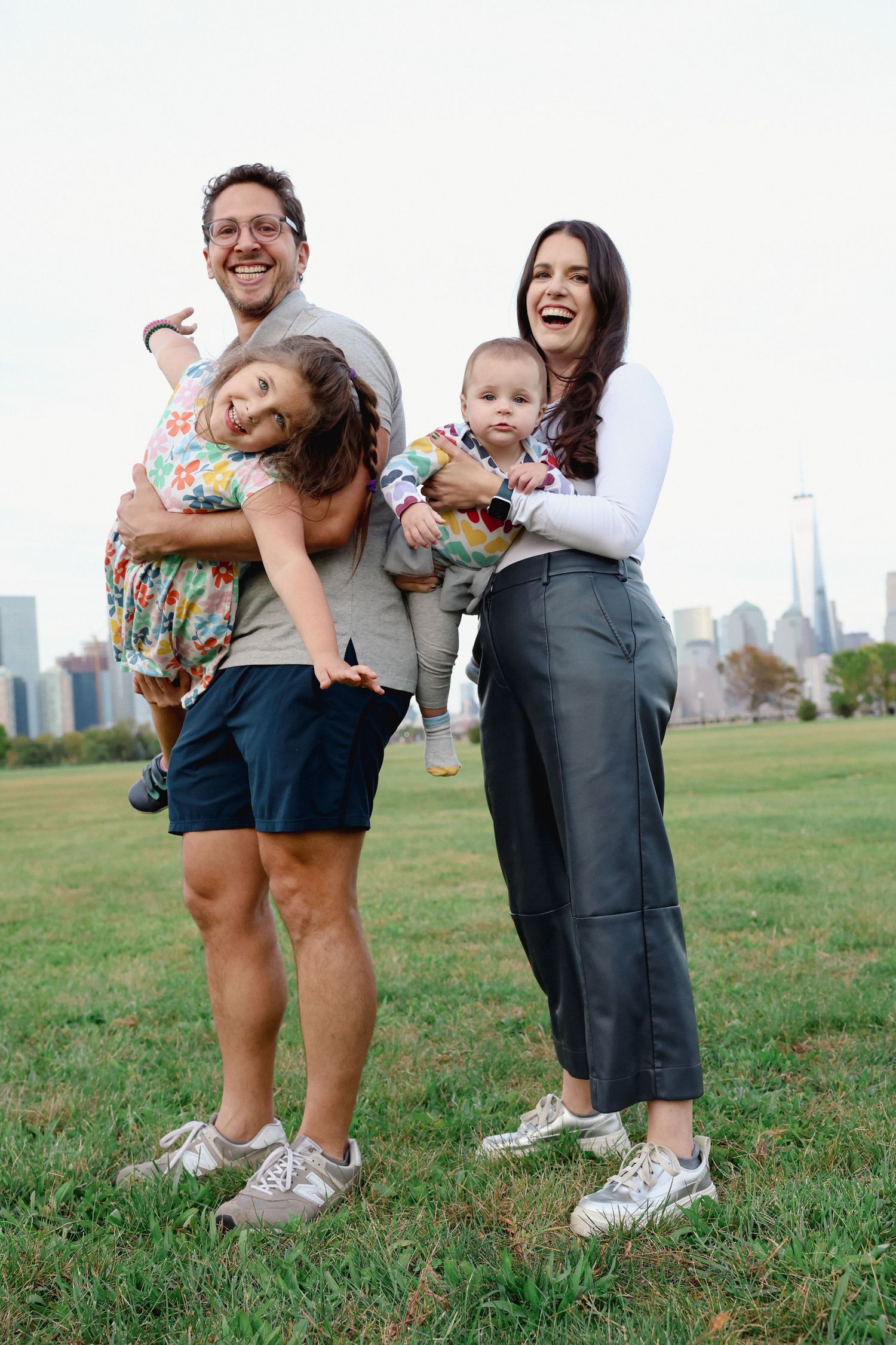 A smiling couple holds two laughing young children on a grassy lawn with a hazy city skyline behind them.