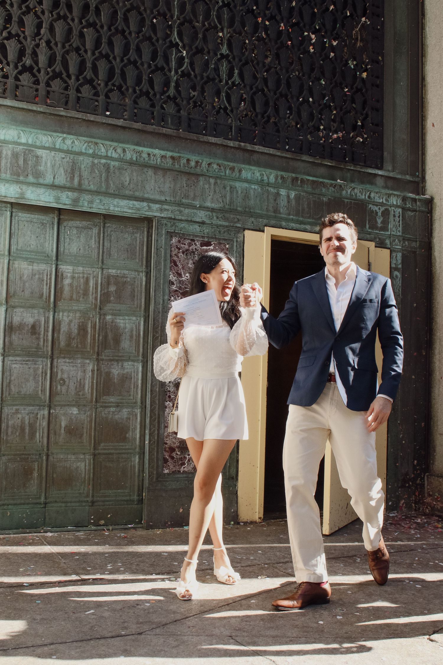 Excited newlyweds walk out of NYC City Hall, bride in white romper holding marriage certificate, laughing joyfully.