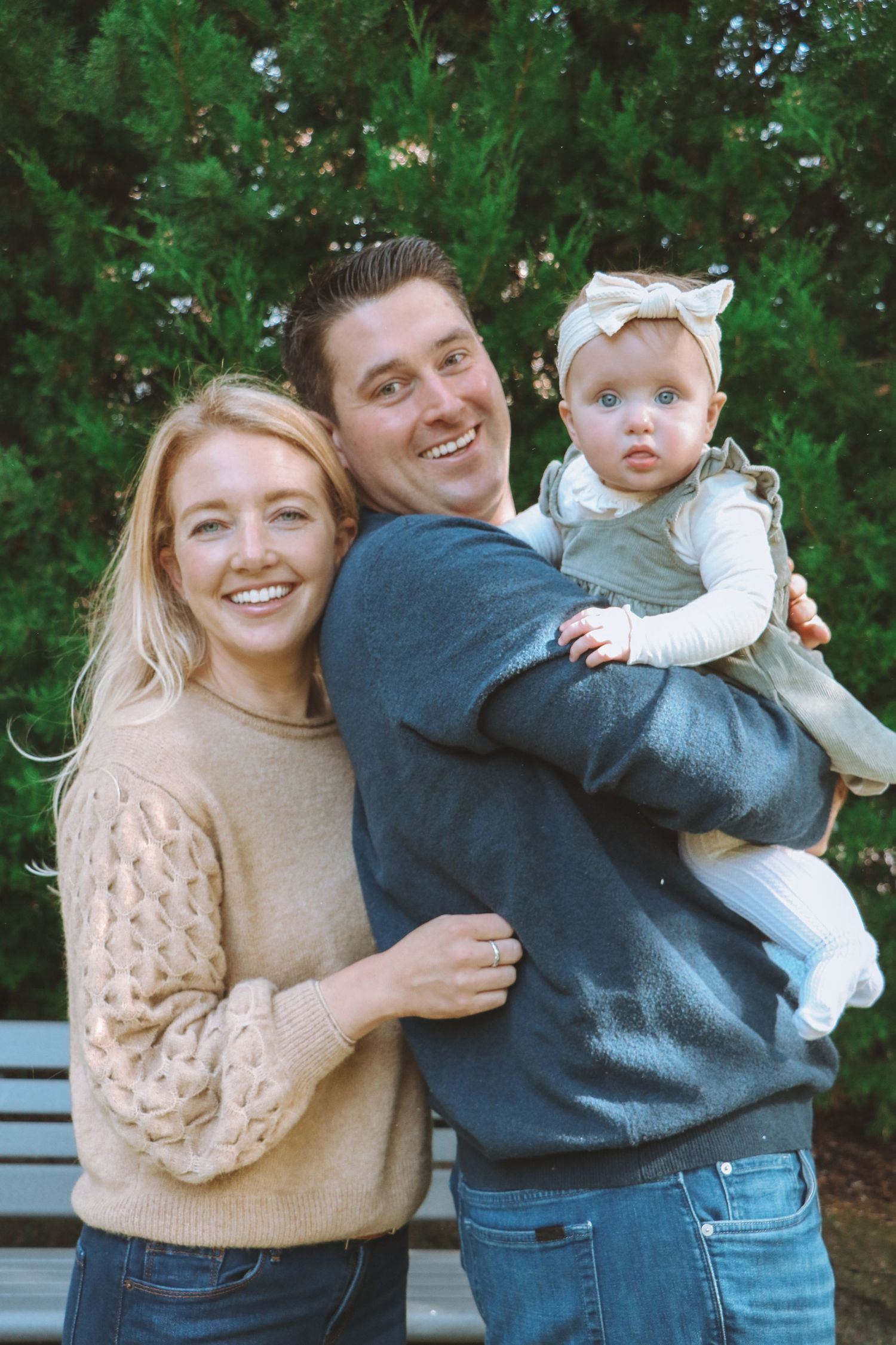 Smiling family of three poses together outdoors with evergreen trees and a park bench behind them.