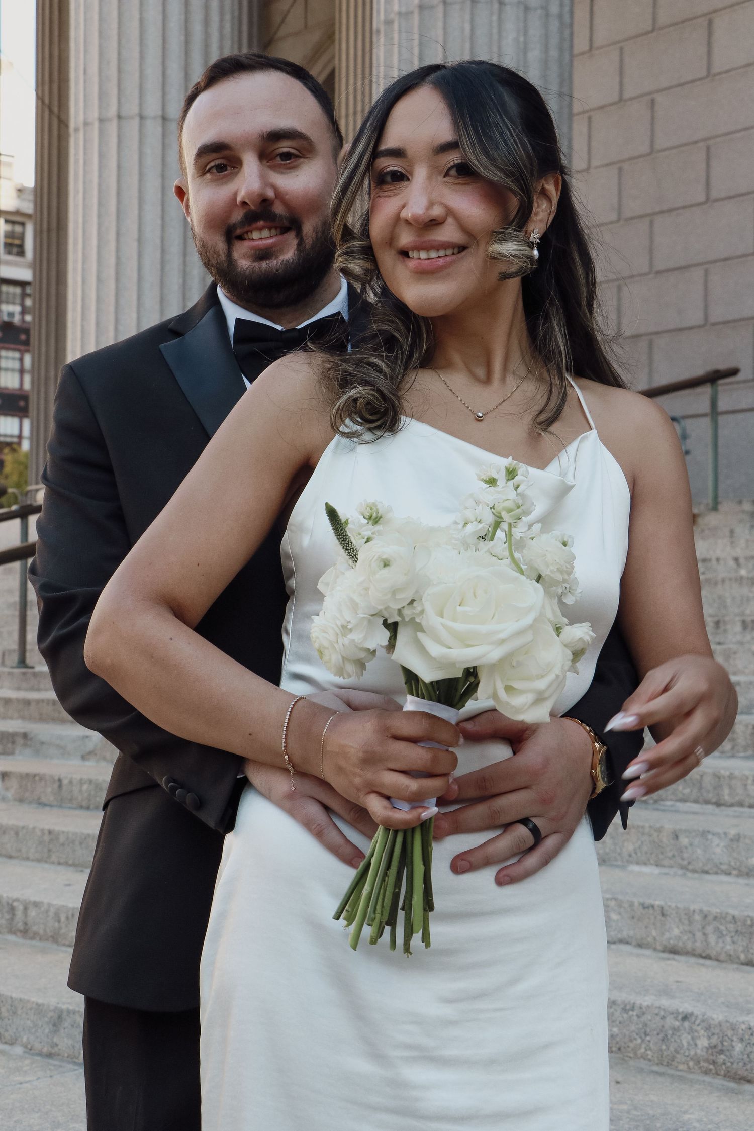 Bride and groom smiling on steps, bride holding white rose bouquet in elegant white gown.