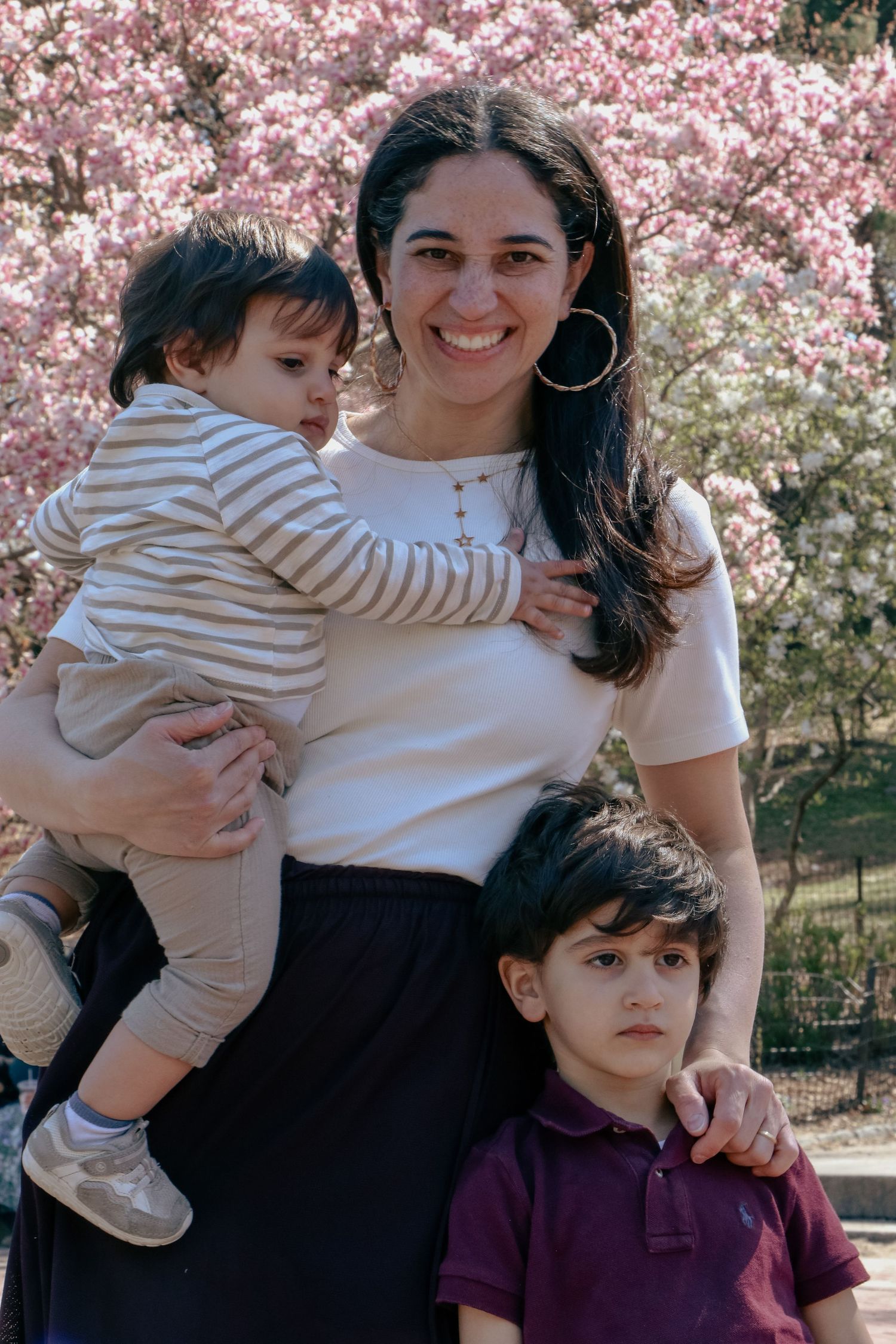 Smiling mother holding toddler with young son beside her in front of pink cherry blossoms.
