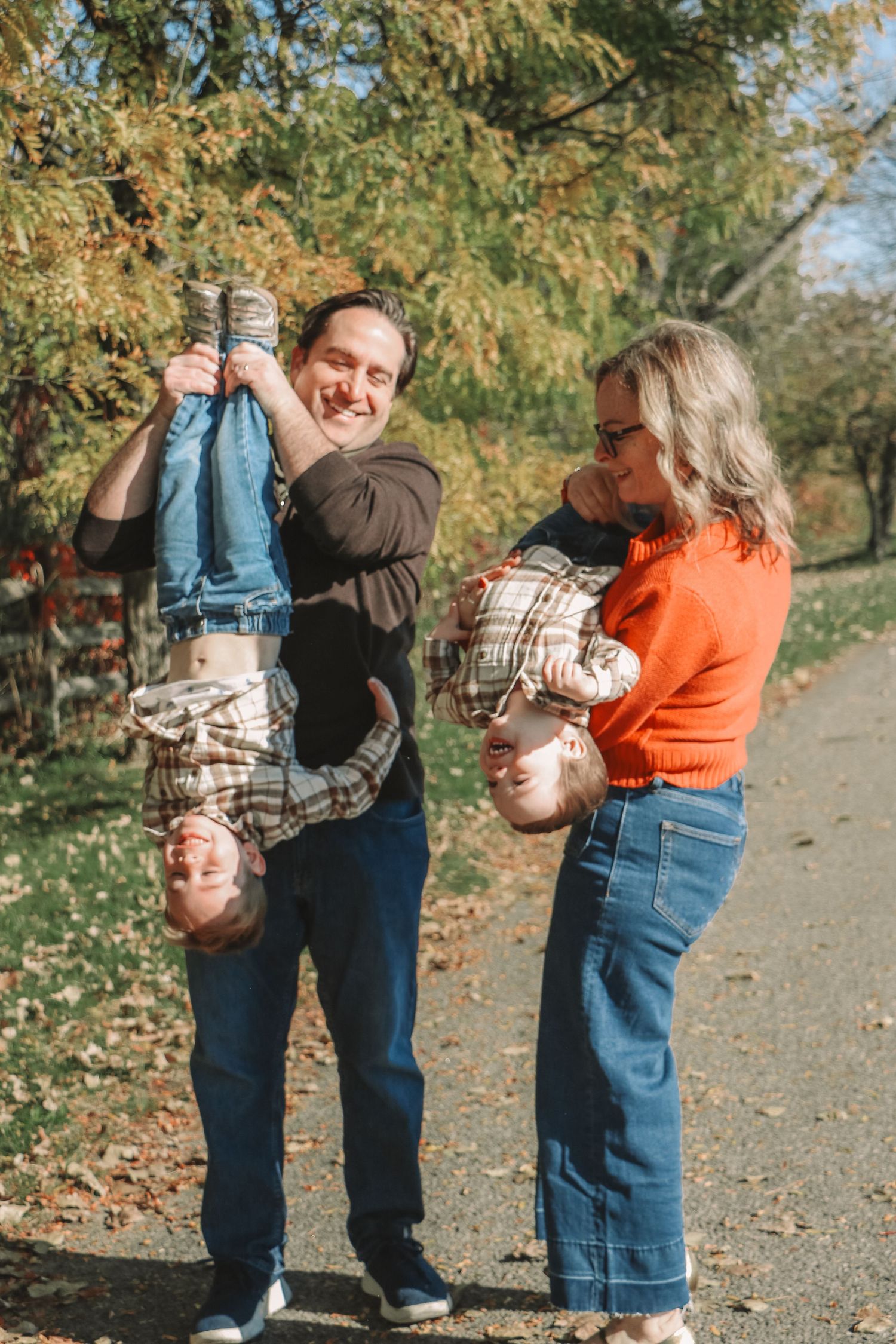 Playful parents holding two young children upside down on a tree-lined path in autumn park.