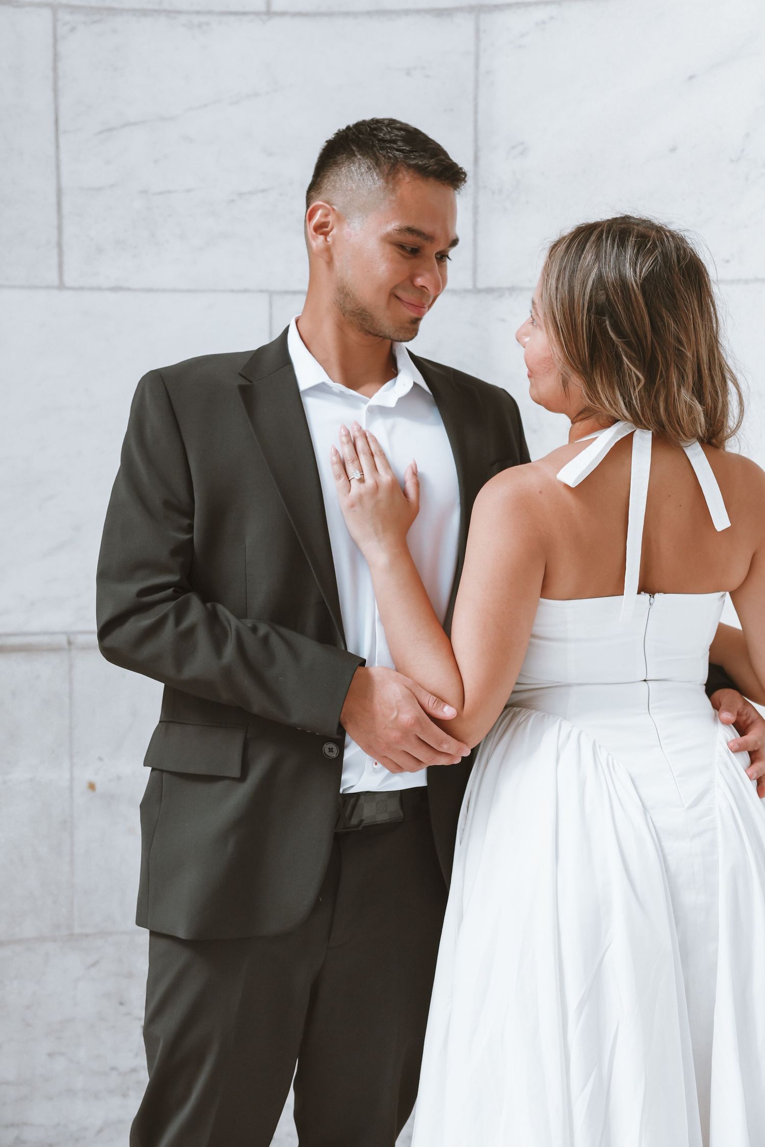 Smiling man in a dark suit gazes at a woman in a white halter dress as she rests her hand on his chest against a marble wall.