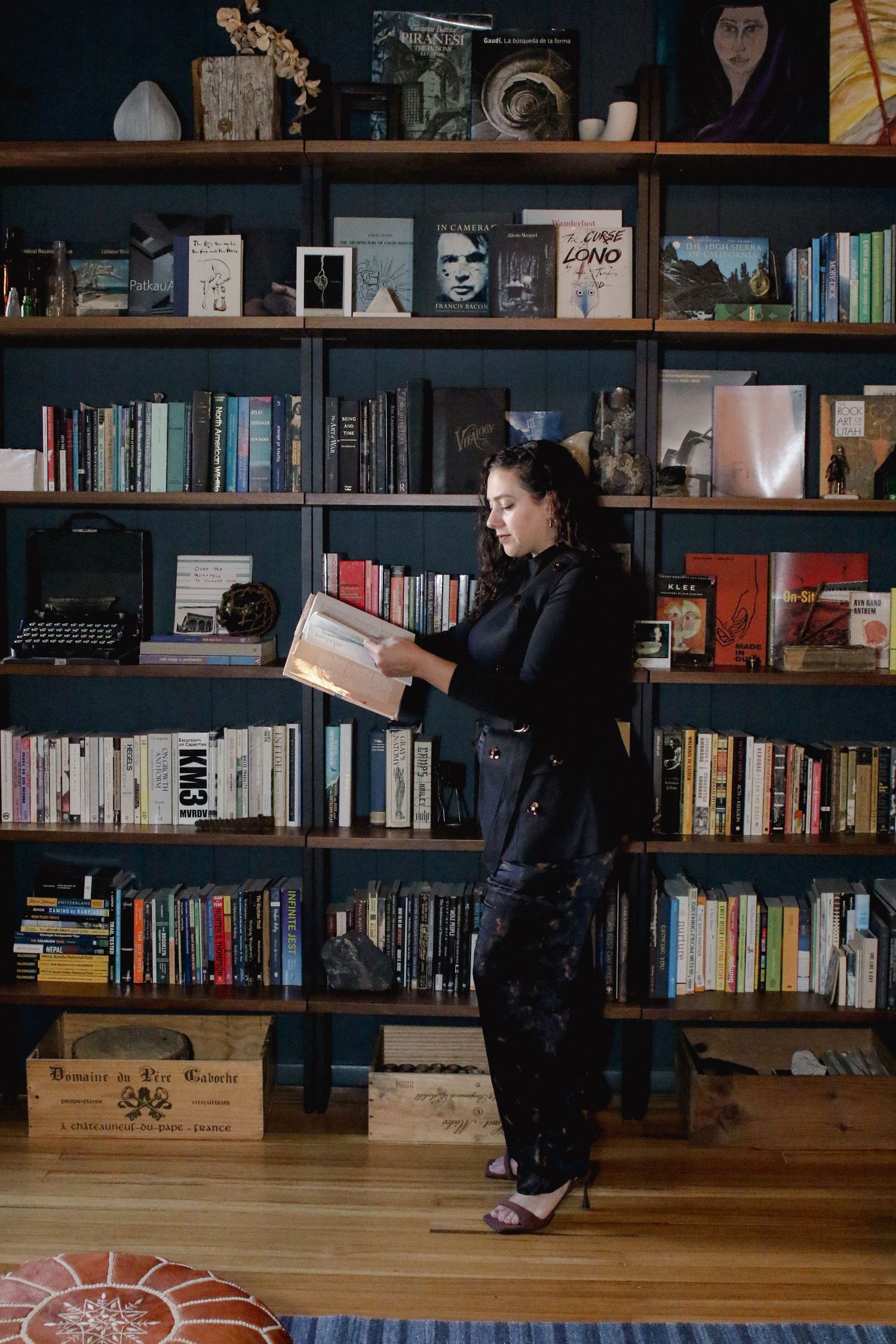 Woman in black outfit reading book while standing in front of large dark bookshelf library.