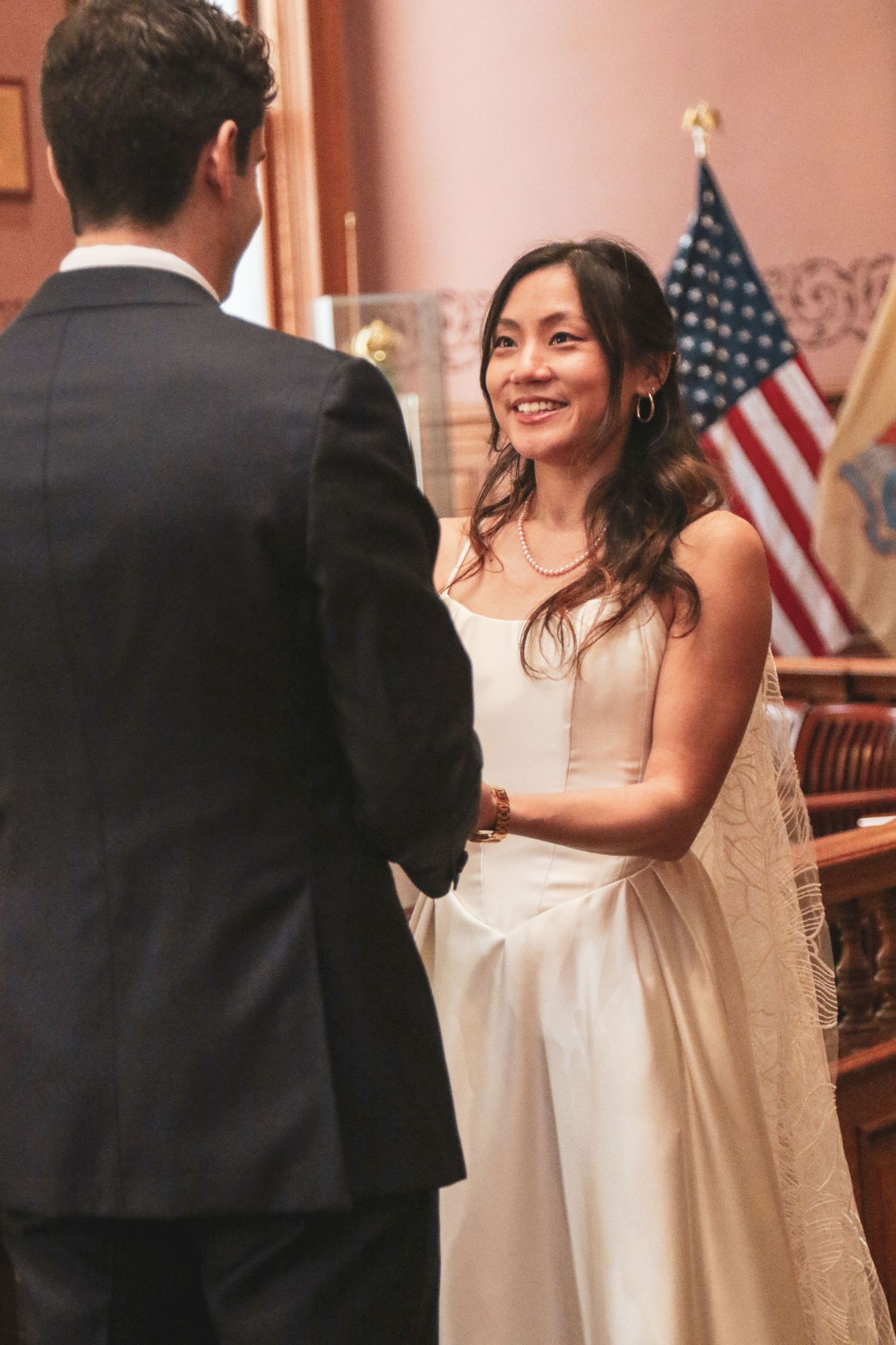 Bride in white gown smiles at groom during civil wedding ceremony with American flag in background.