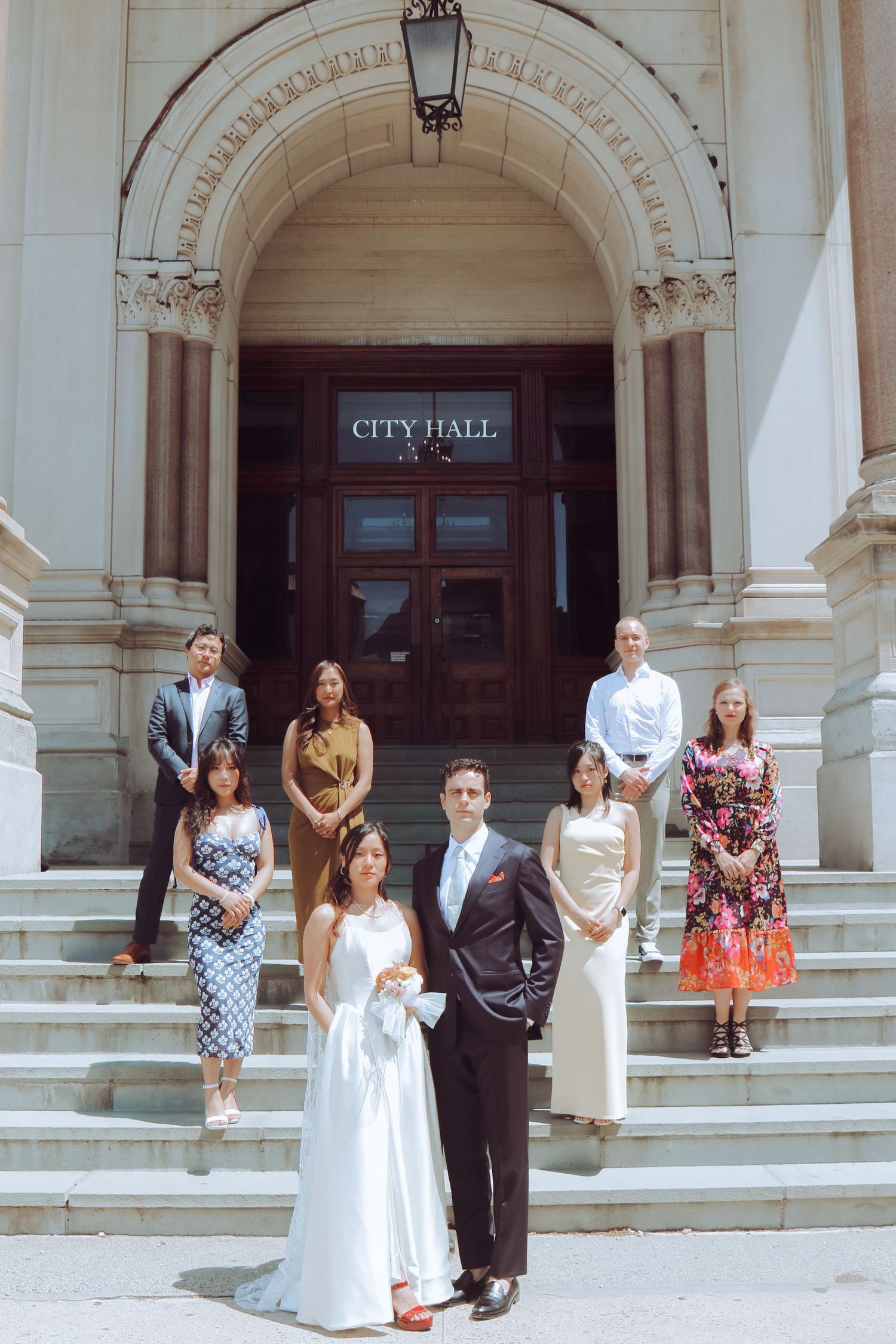 A wedding couple poses with guests on the steps of a historic City Hall building on a sunny day.