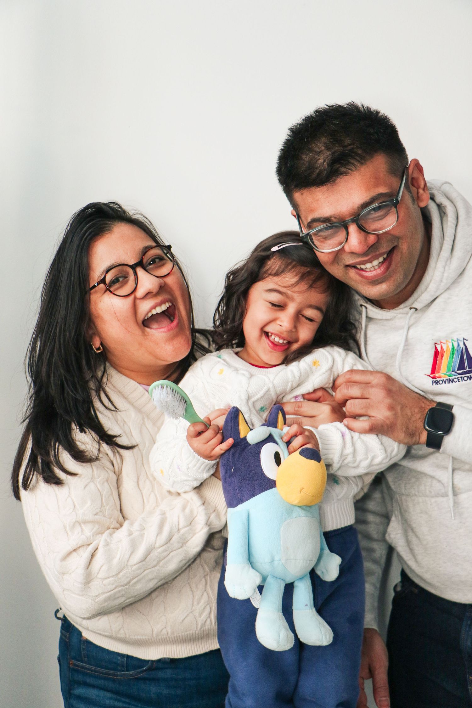 Laughing family of three with parents hugging their child who holds a Bluey plush toy in a white studio.