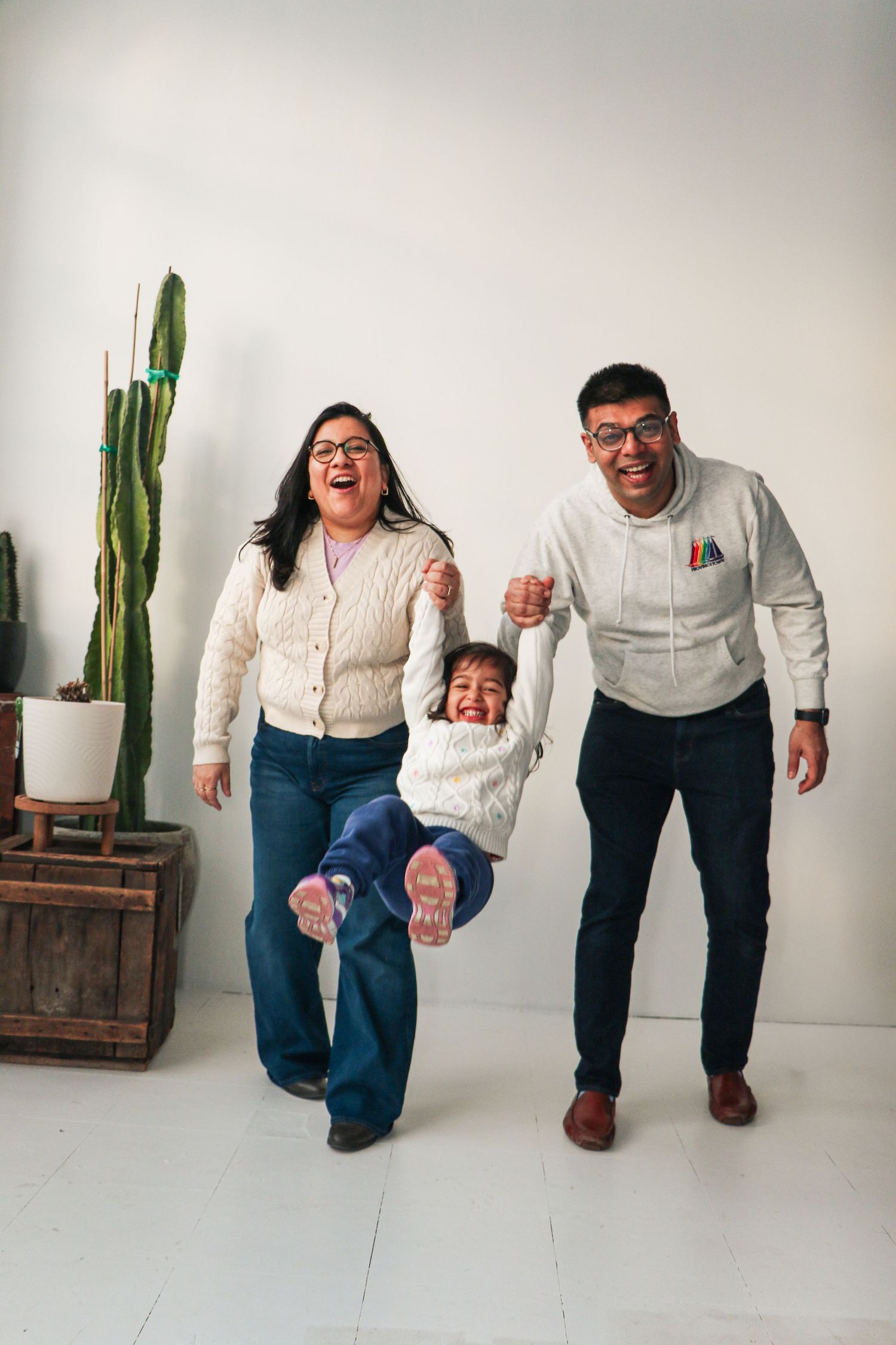 Happy family with two parents swinging their laughing child between them in a bright white studio with a cactus.