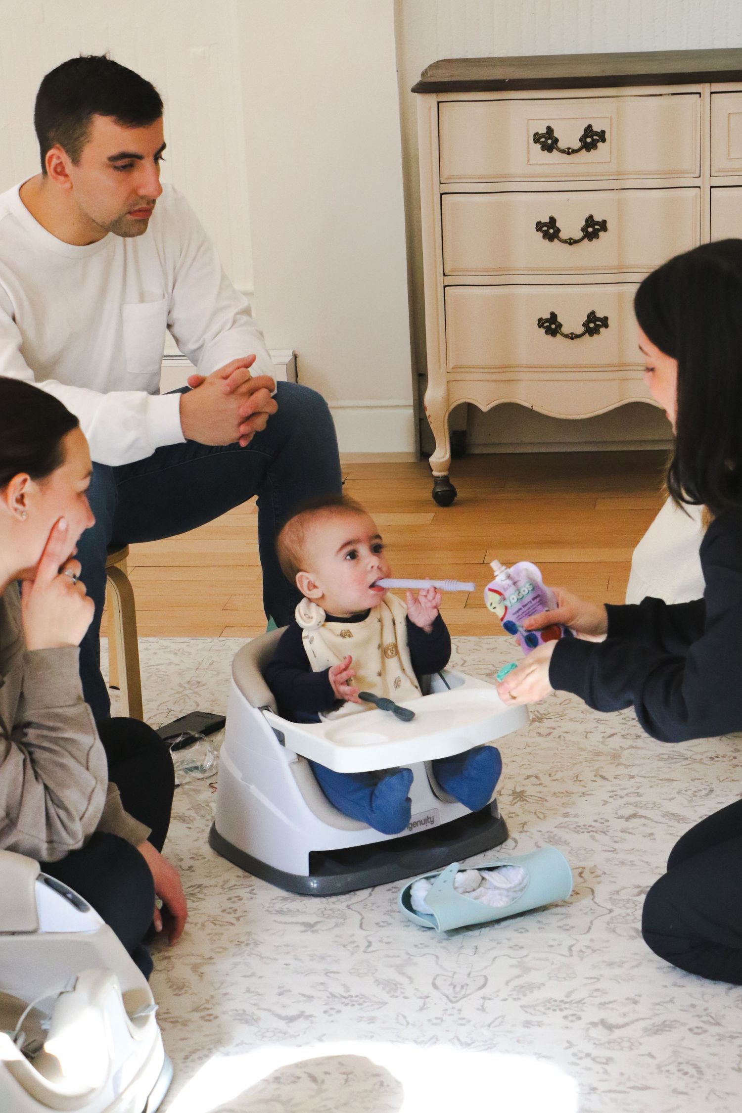 Parents and a woman gathered around a baby seated in a floor booster seat being fed with a spoon.