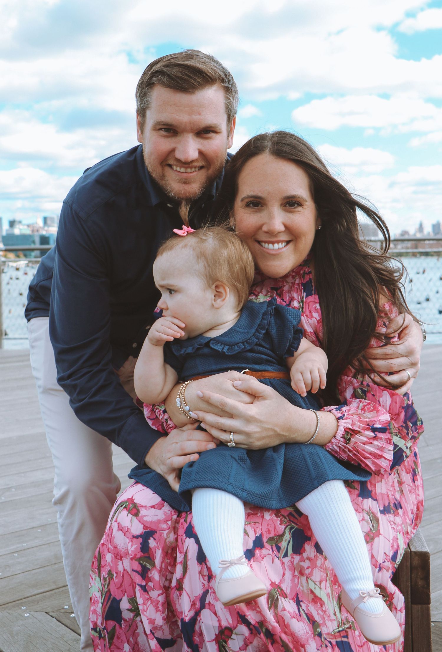 Smiling family portrait with parents and baby girl on outdoor deck with city skyline.