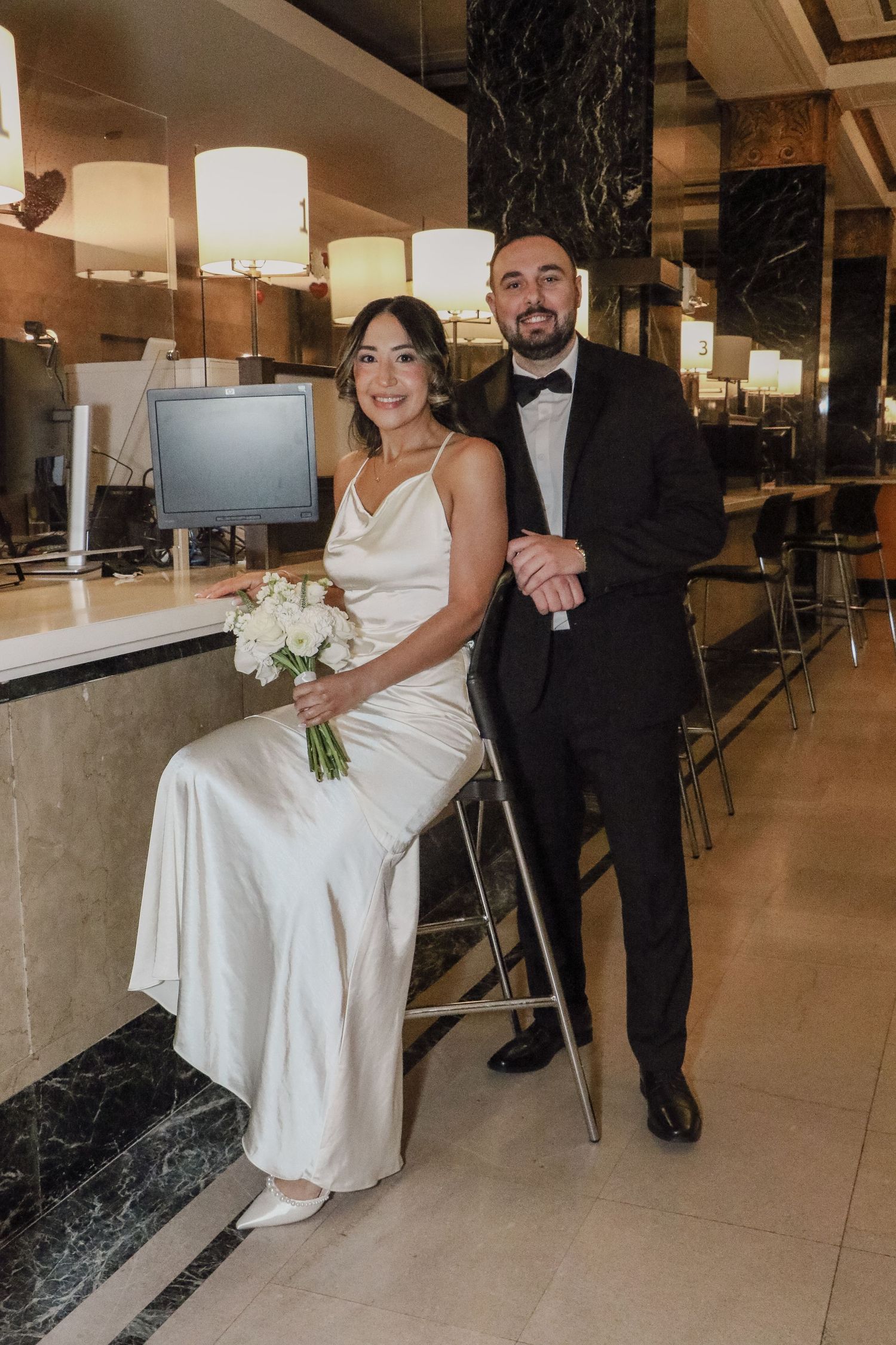 Elegant bride in white satin slip dress with bouquet sits at bar beside groom in black tuxedo at NYC wedding.