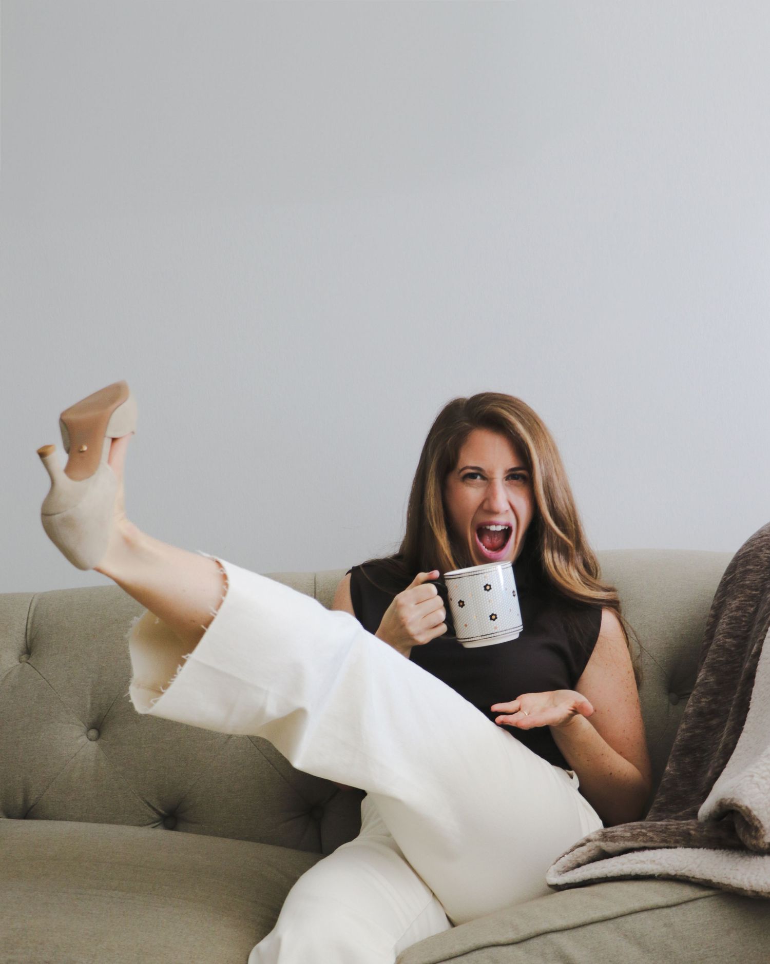 Excited woman in black top and white pants holding a mug while lounging on a gray sofa.