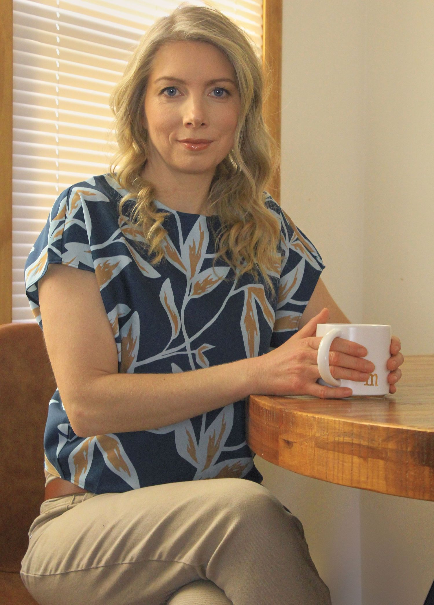 Blonde woman in navy floral blouse holding white coffee mug while seated at wooden table.