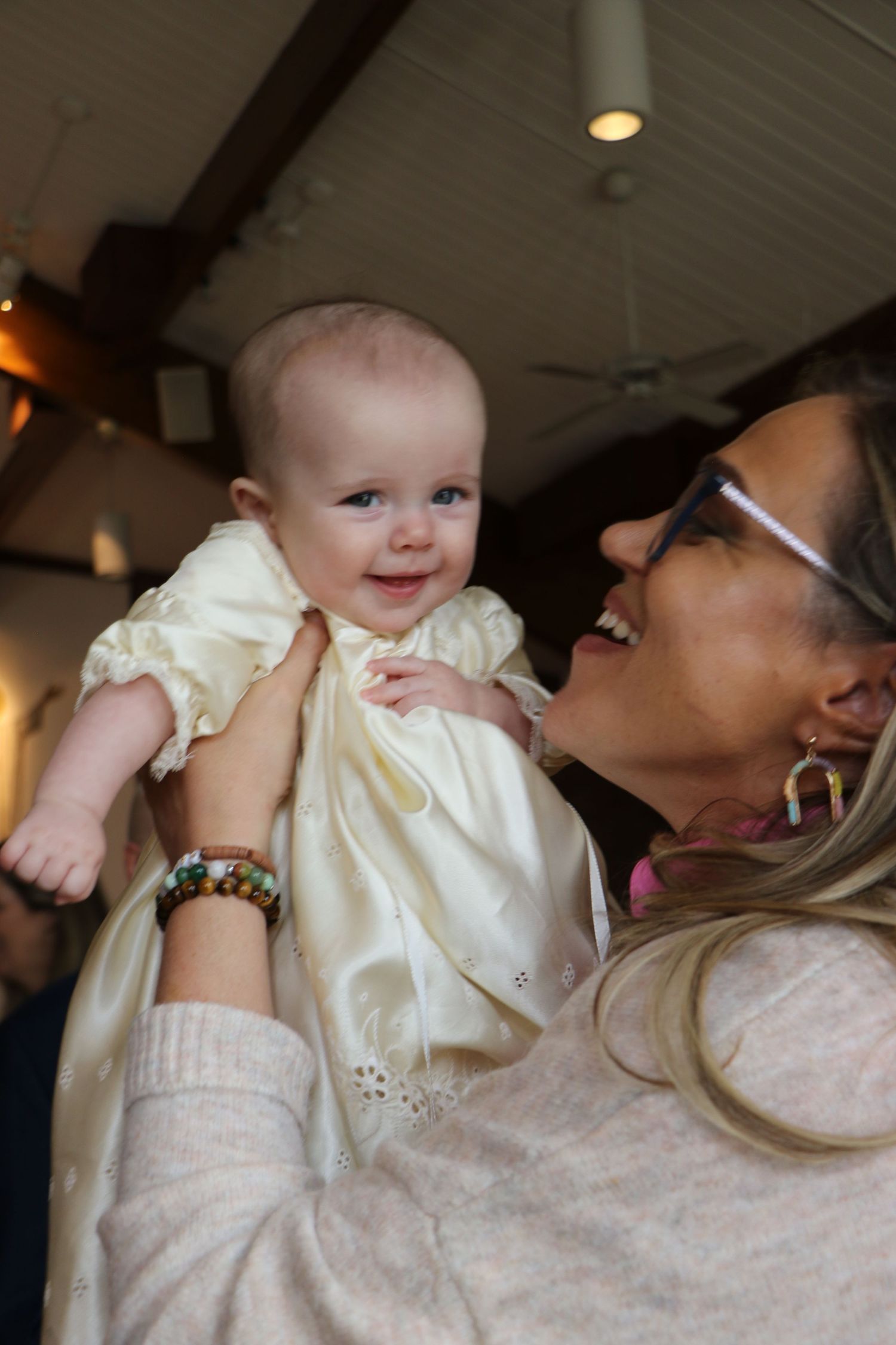 A woman lifts a smiling baby in a cream christening gown joyfully after a baptism ceremony.