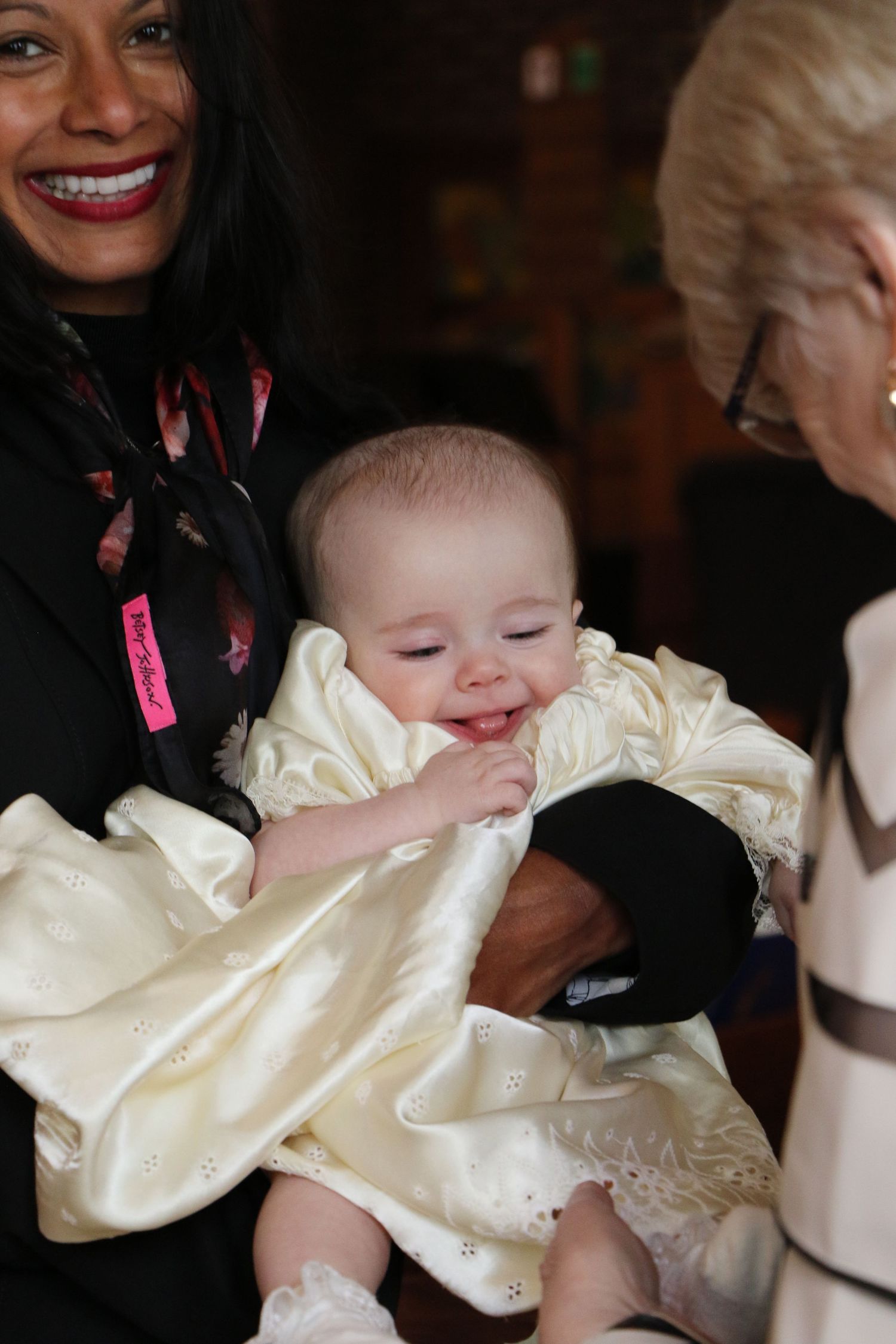 Smiling woman holding a baby in a white christening gown at a baptism.