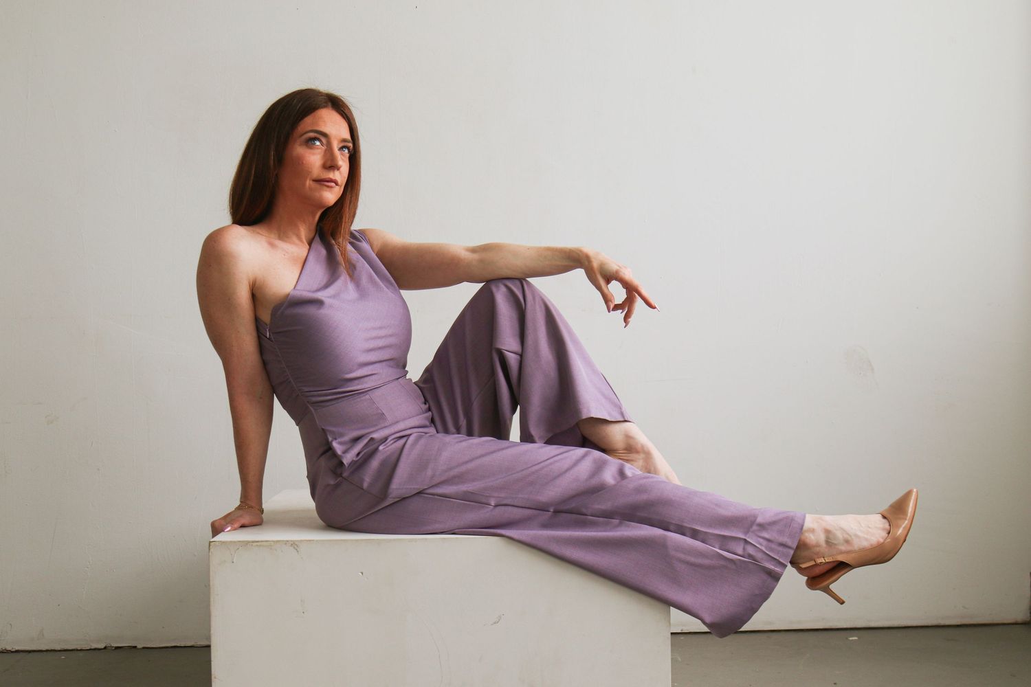 Woman in lavender halter jumpsuit and nude heels poses elegantly on white cube against light gray background.