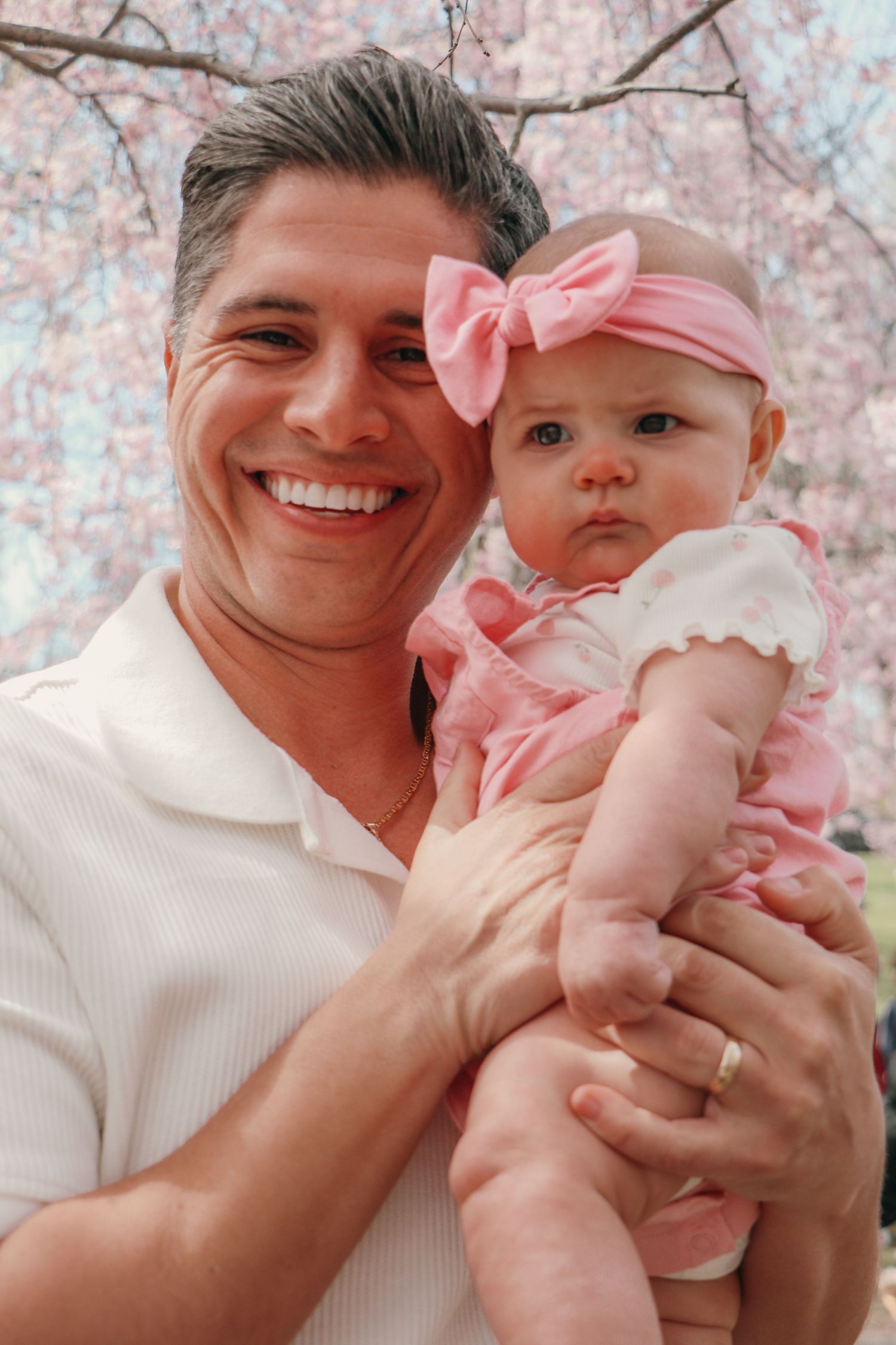 Smiling father holds baby girl in pink bow and dress under cherry blossom tree.