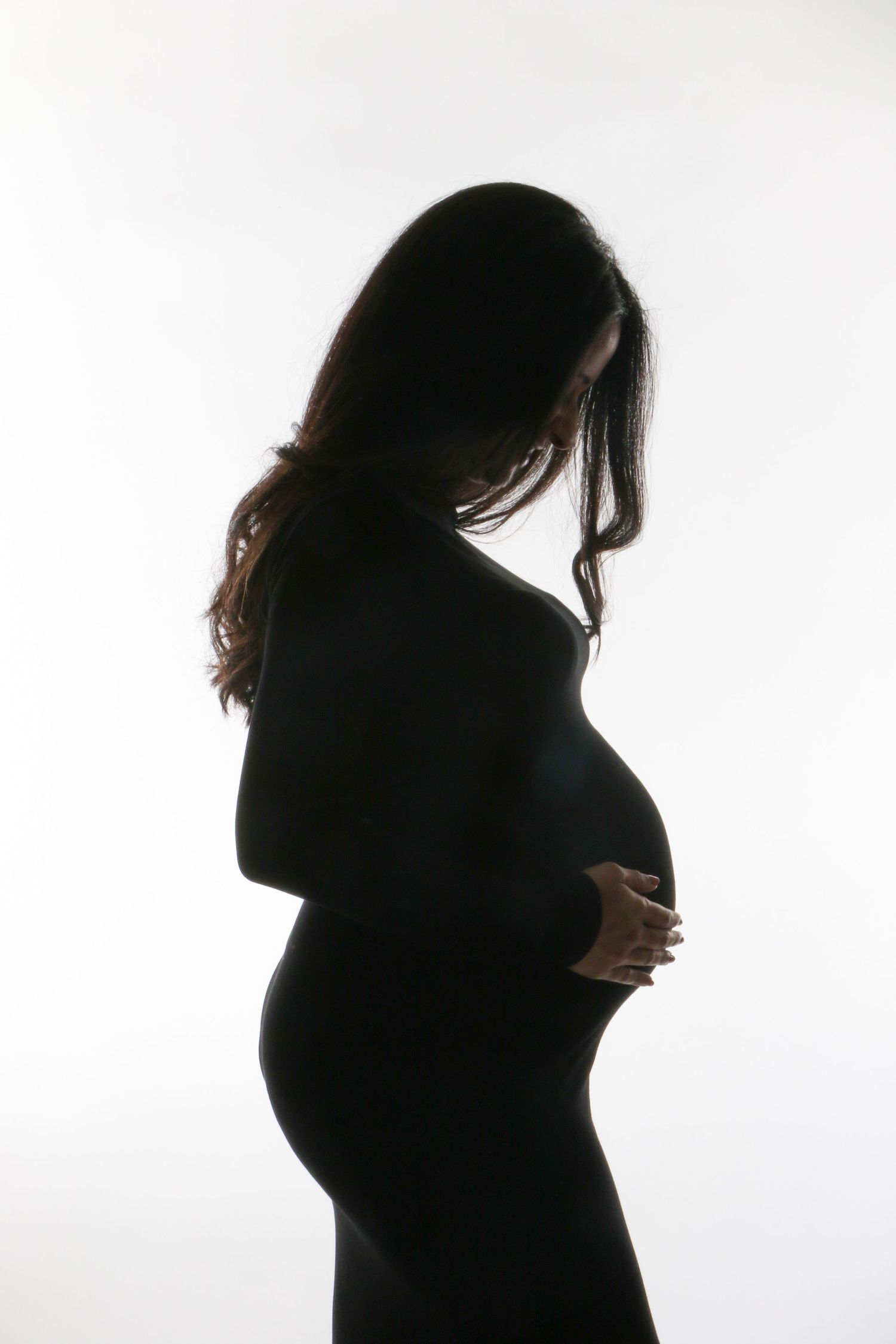 Dark silhouette of pregnant woman gently holding her belly against white studio background.