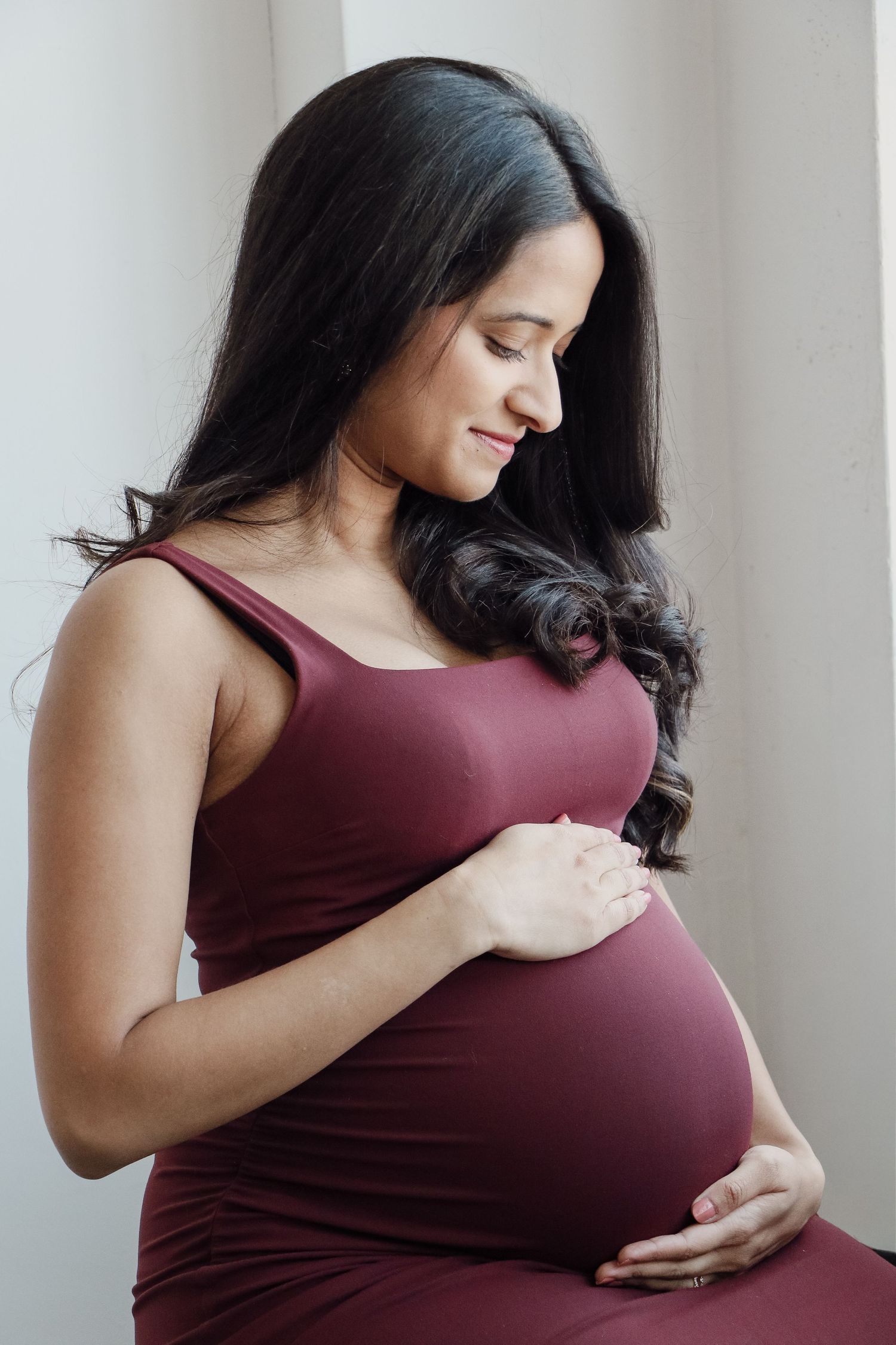 Pregnant woman in dark red dress lovingly cradles her baby bump in soft studio lighting.