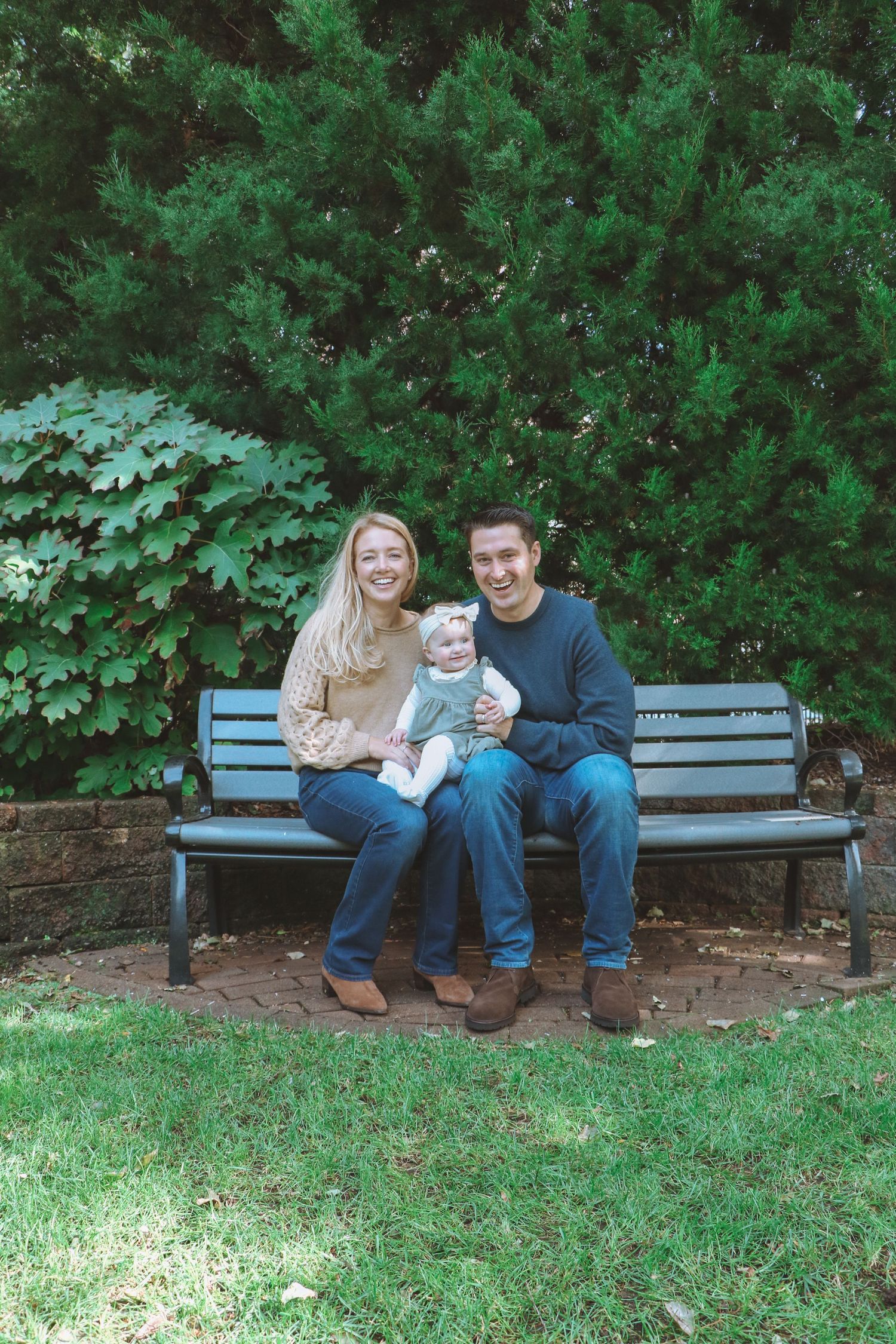 Smiling couple with baby sitting on bench in lush green garden setting.