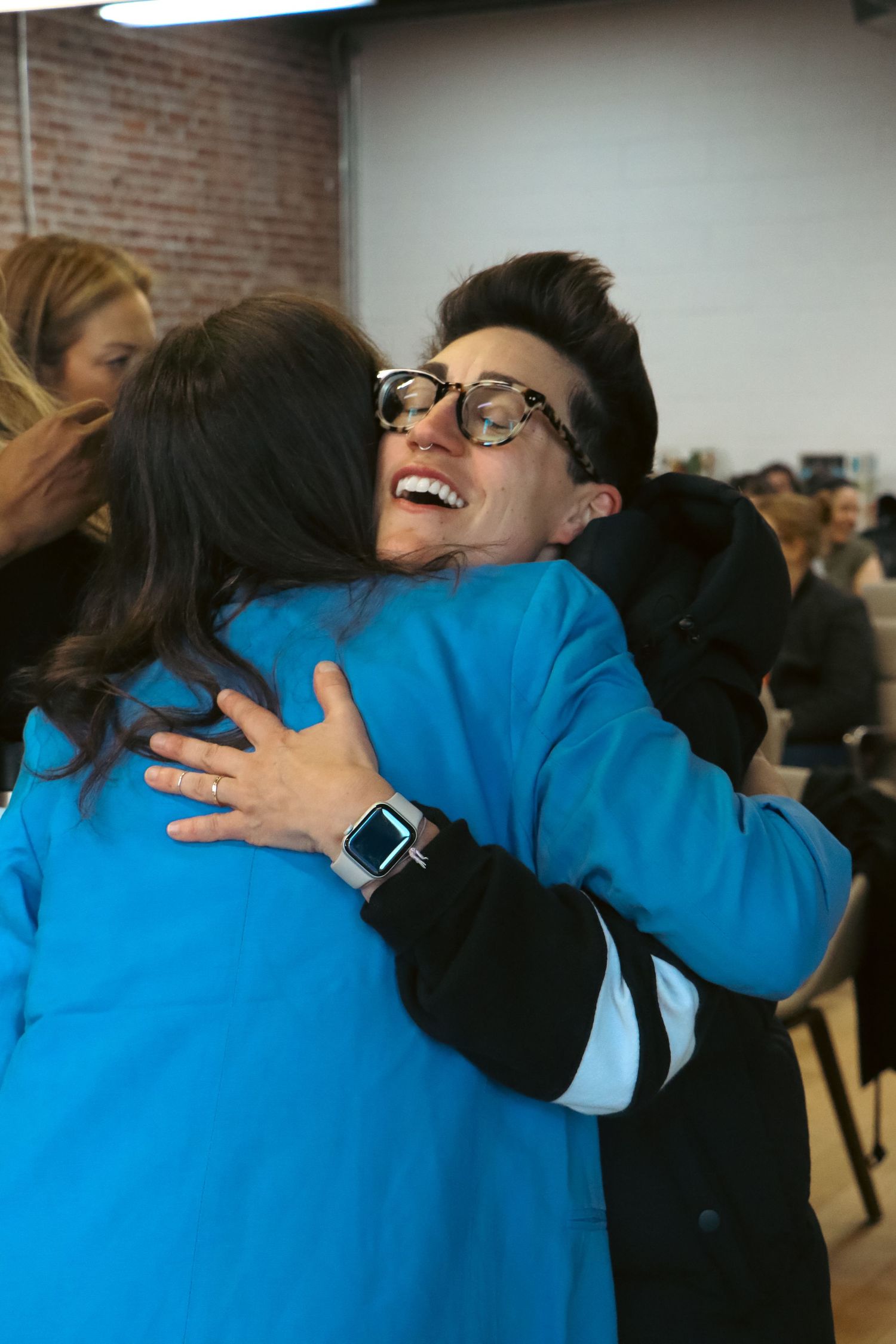 Two women sharing a warm hug at an indoor event, one wearing blue jacket and glasses.