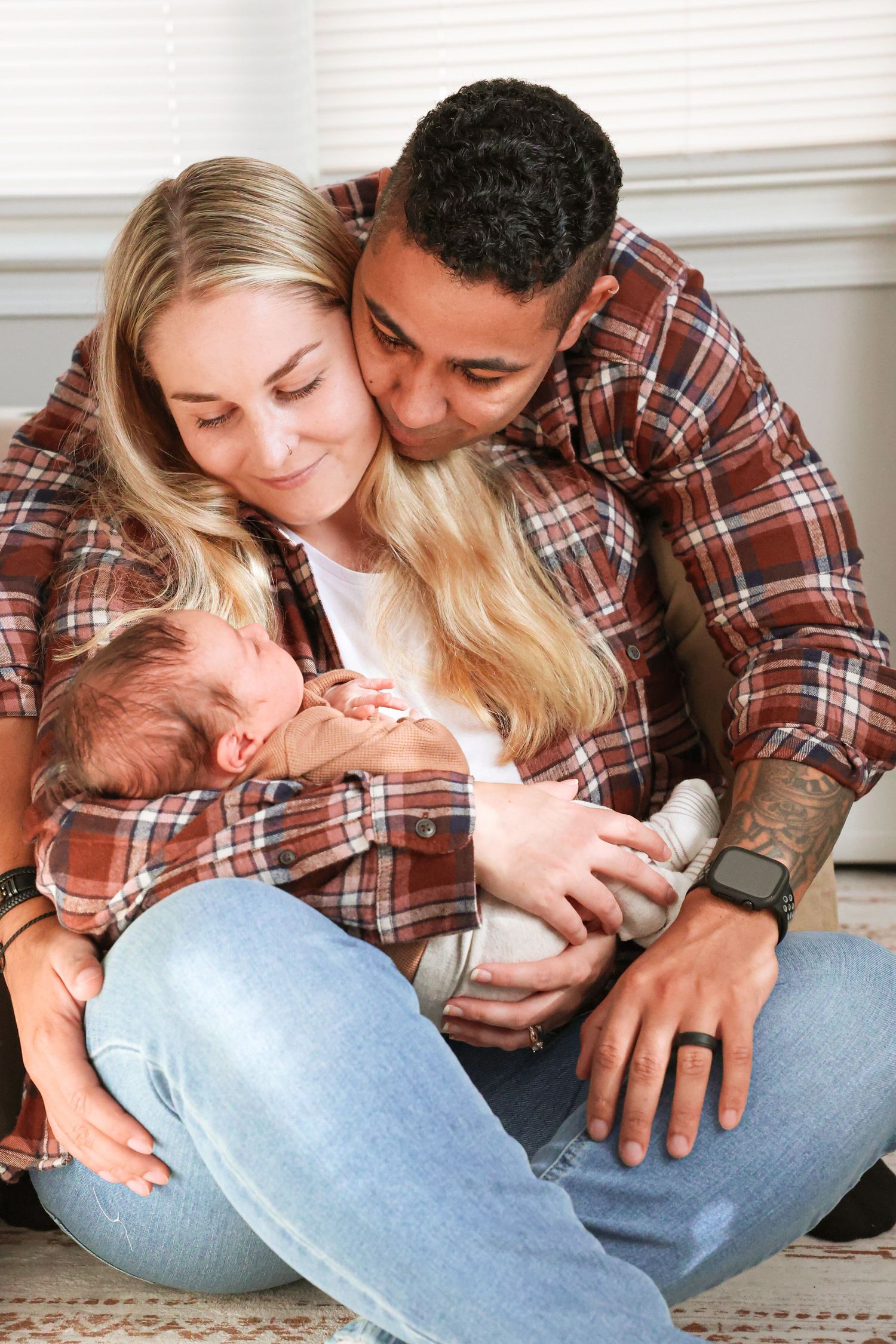 A smiling woman and a man in matching plaid flannel shirts sit close on a rug, gently cuddling their sleeping newborn baby.