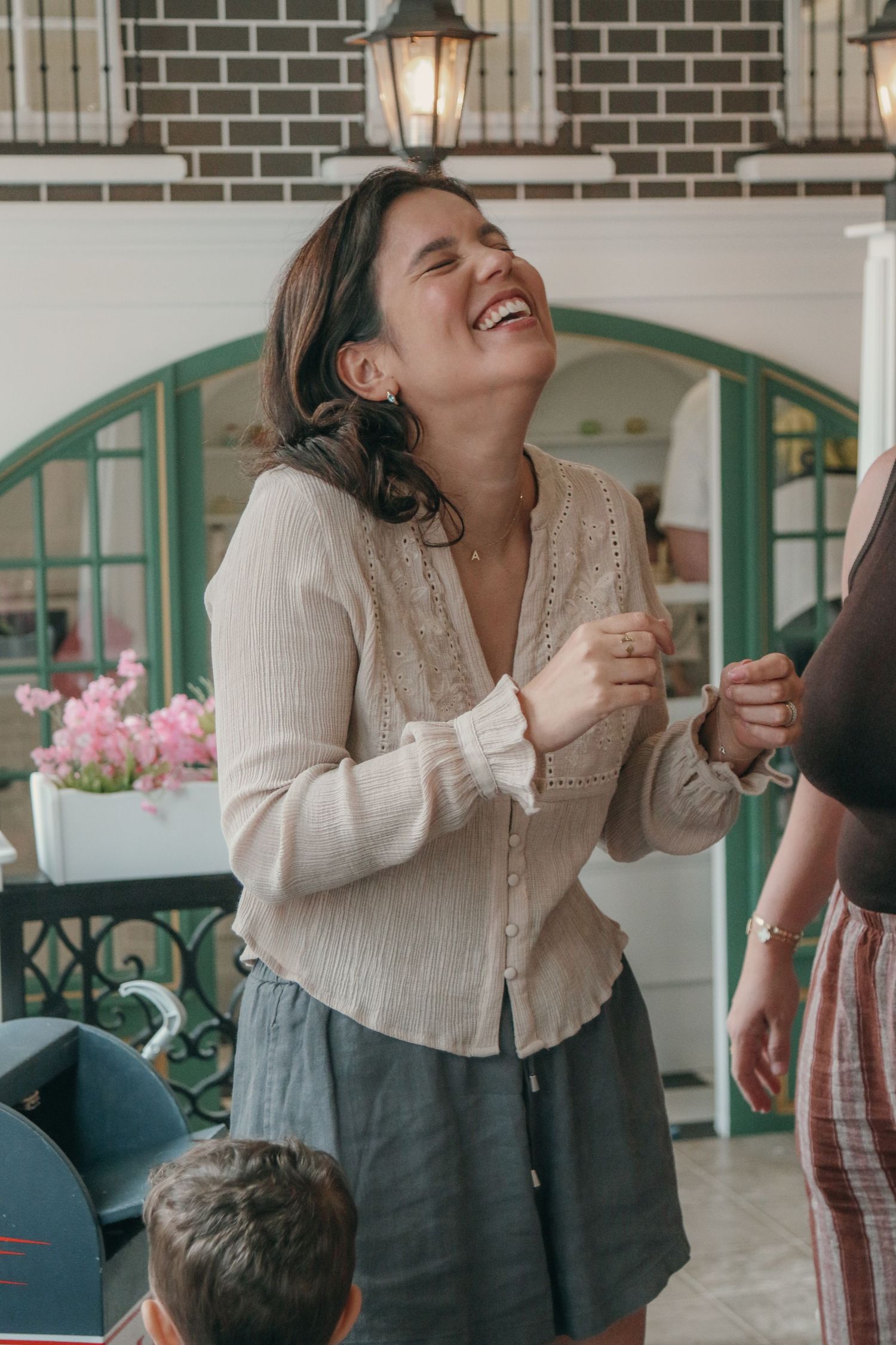 A laughing woman in a cream knit cardigan and gray skirt stands near pink flowers in a bright cafe with green arched windows.
