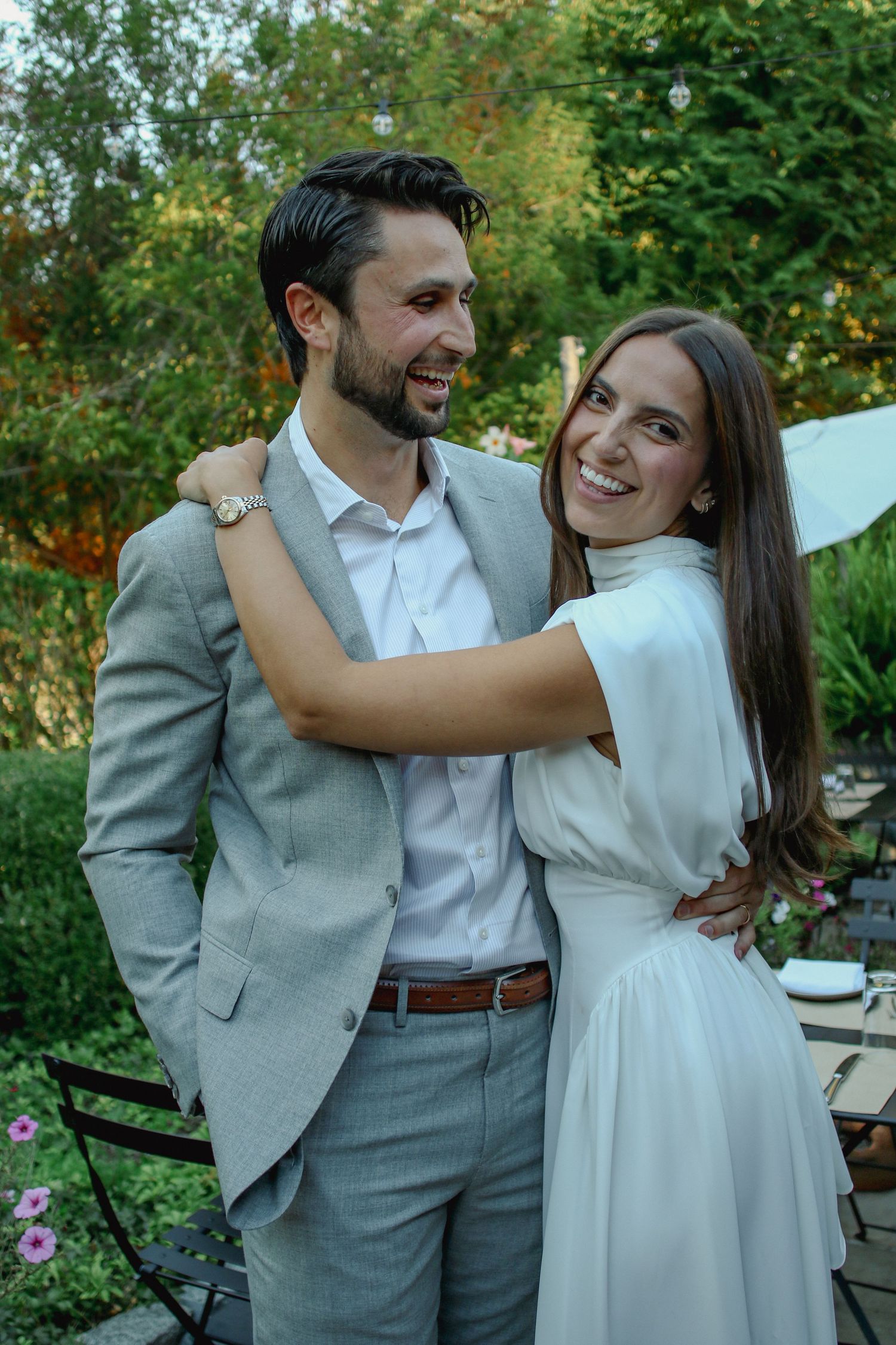 Bride in white dress embraces groom in grey suit while smiling in a lush garden setting.