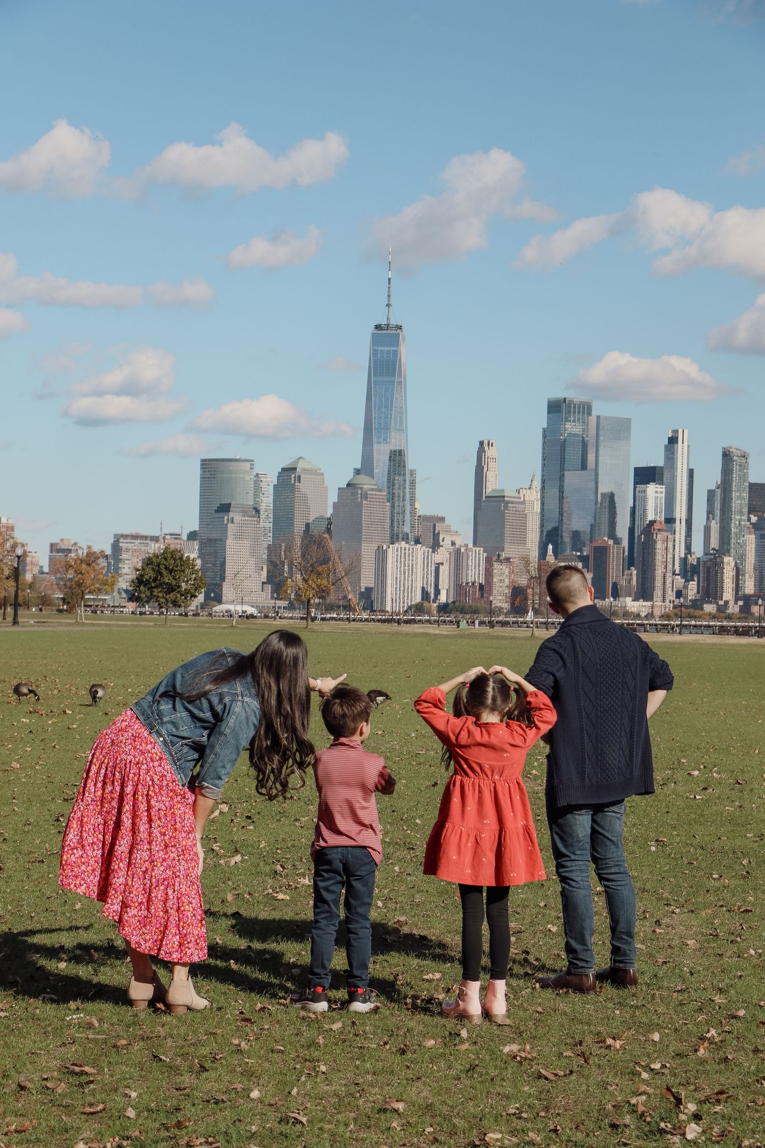 Family of four stands on grass admiring the New York City skyline with One World Trade Center on a sunny day.