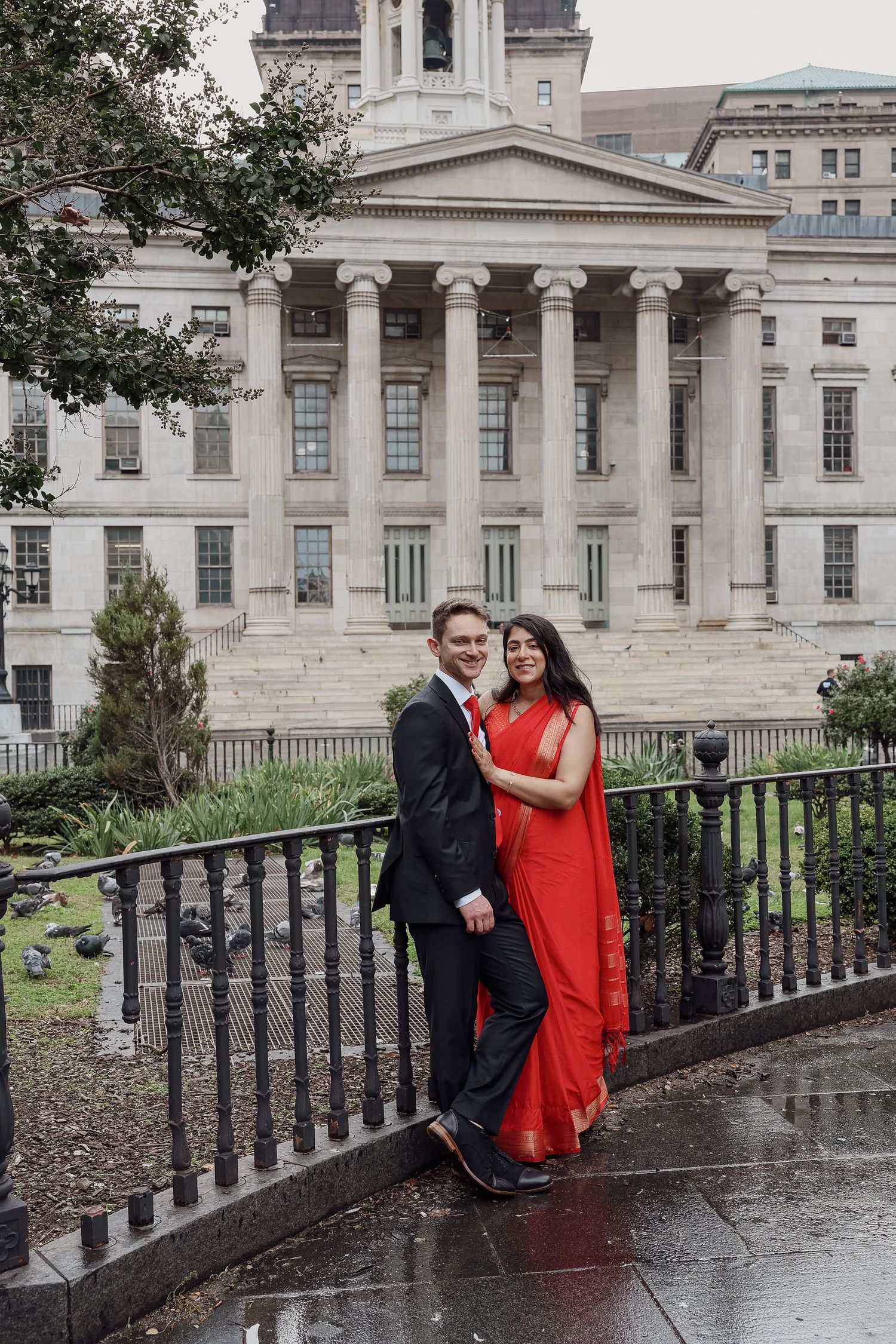 Couple in a black suit and red sari pose together beside a black iron fence in front of a columned neoclassical building on a rainy day.
