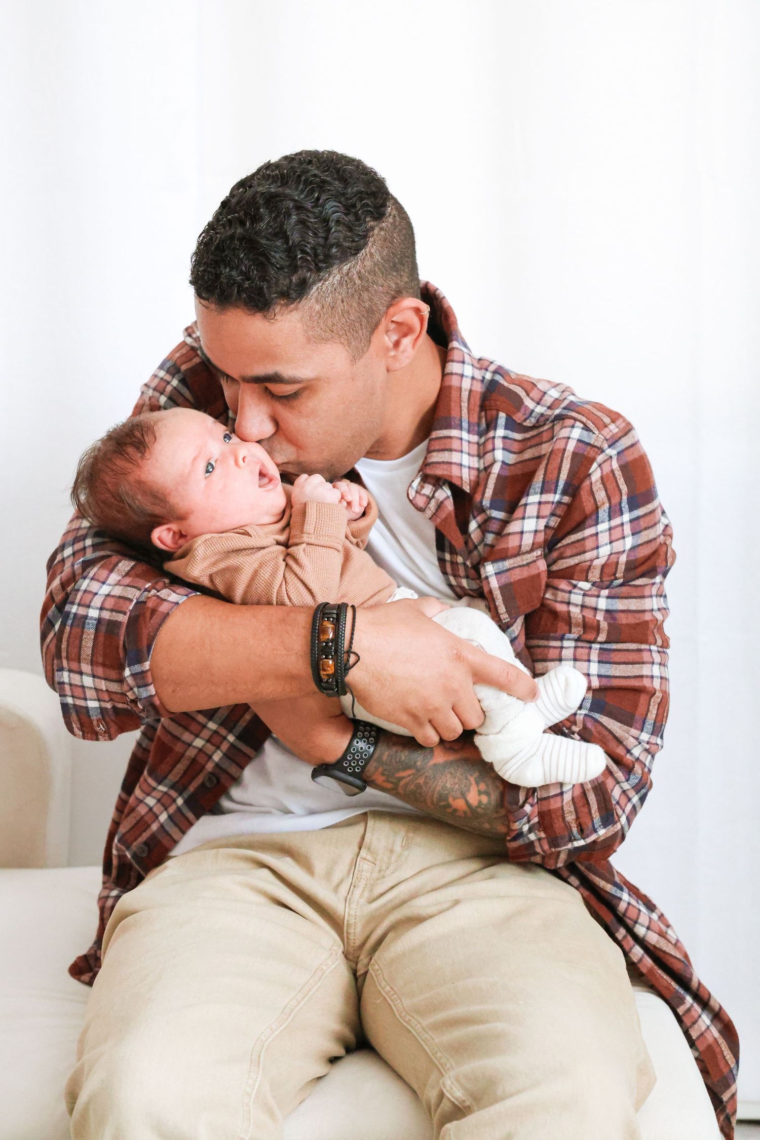 A man in a plaid flannel shirt and khaki pants sits on a white couch, tenderly kissing the head of a baby in a tan outfit.